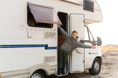 A person standing in the open doorway of a white camper van in a desert landscape, smiling with arms outstretched.