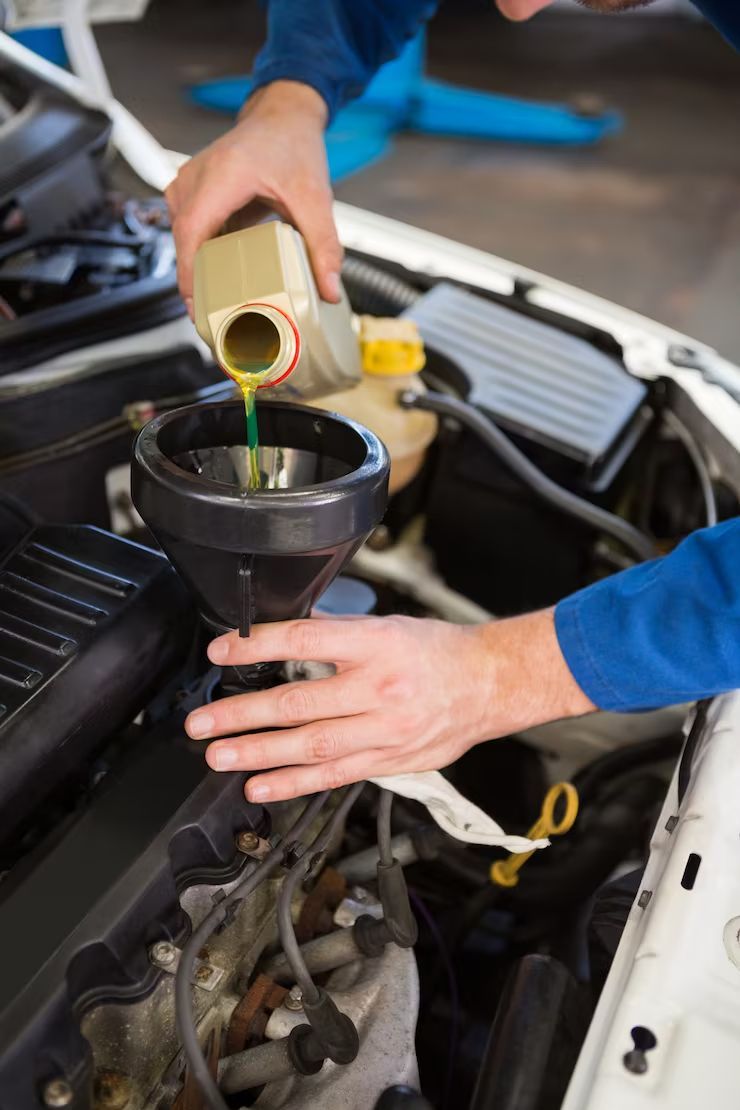 A mechanic in a blue uniform pours bright green engine fluid into a car engine through a black funnel.