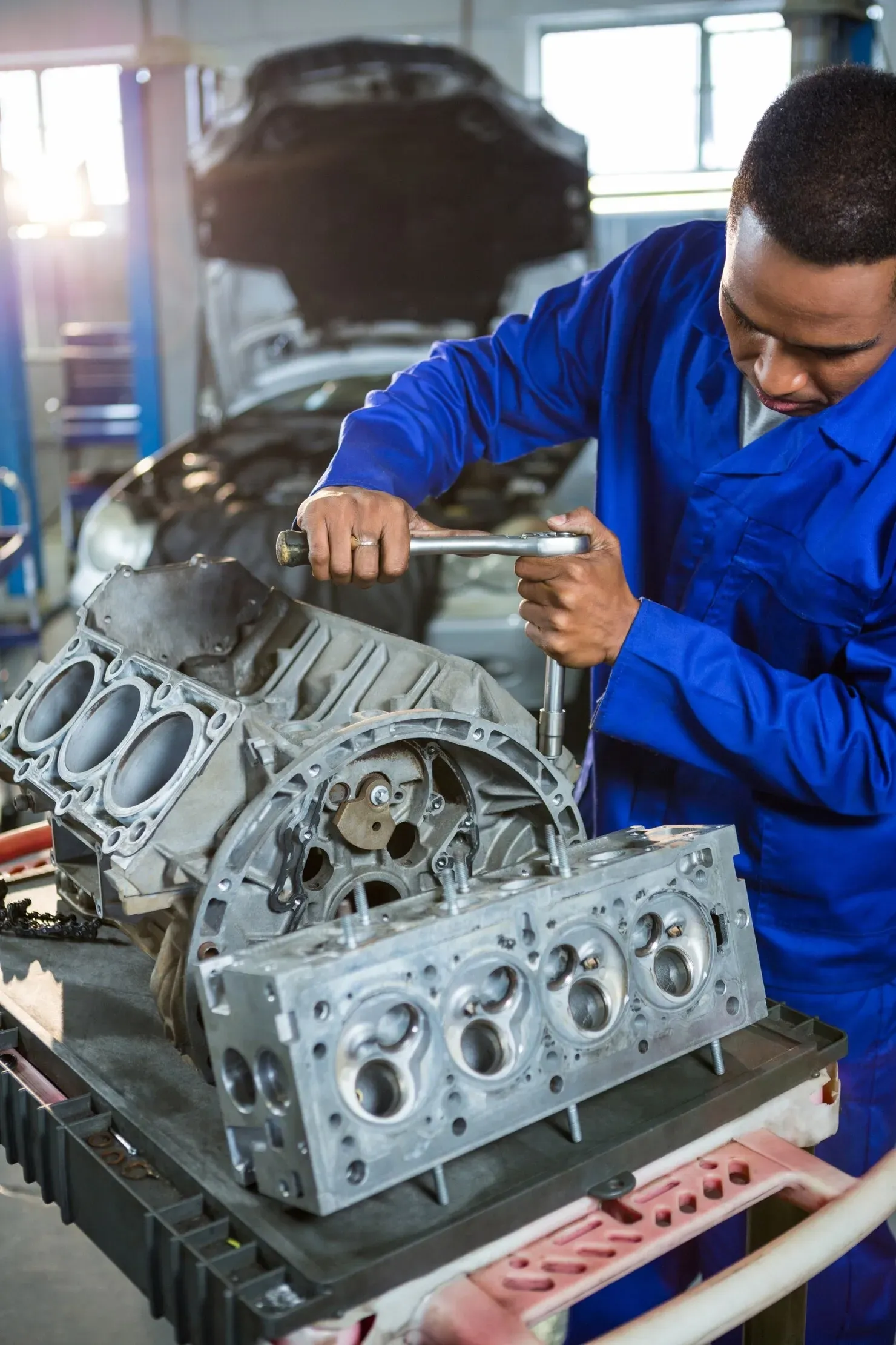 A person in blue coveralls uses a socket wrench to work on a disassembled vehicle engine in a workshop.