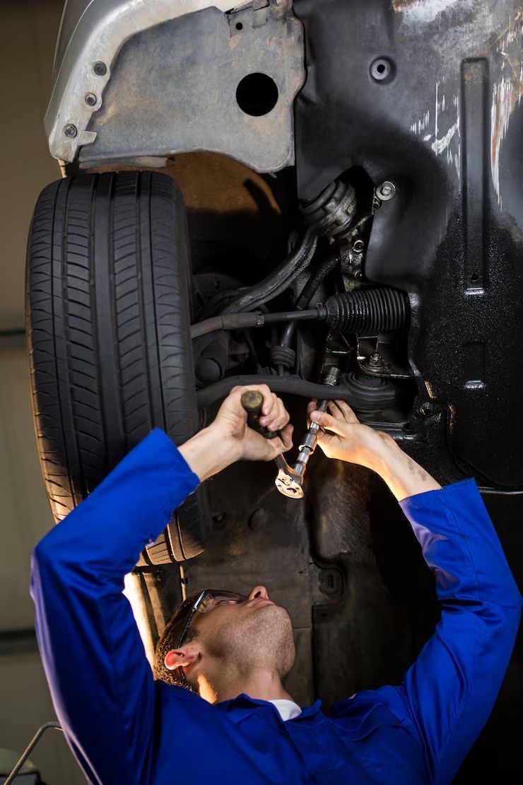 A mechanic in blue coveralls uses a flashlight to inspect the undercarriage of a car raised in a garage.