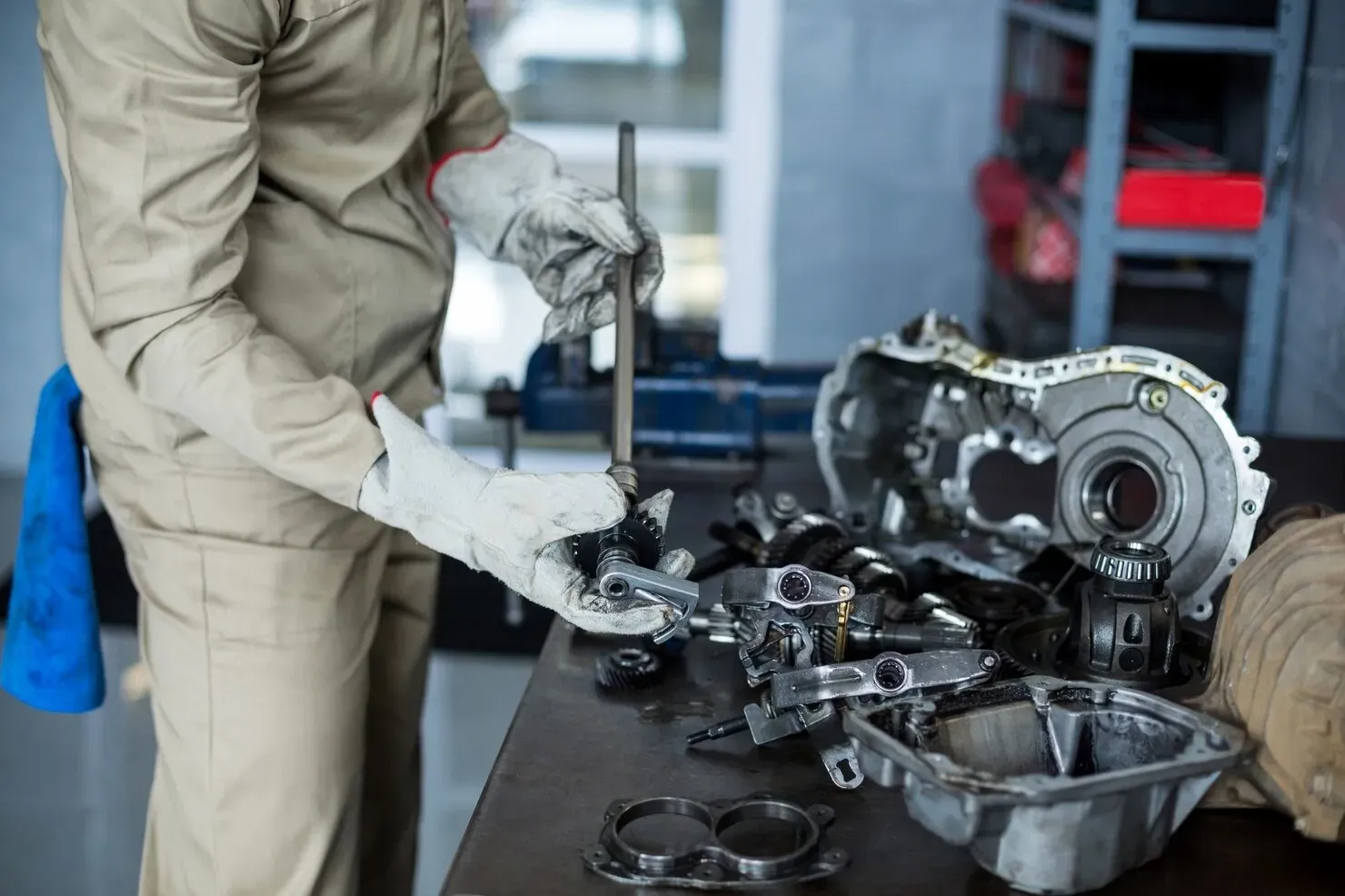 A person in tan work coveralls and gloves assembles automotive parts on a workshop table.