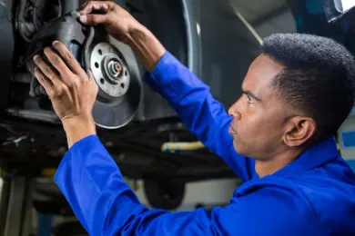 A mechanic wearing a blue uniform inspects a car’s brake system in a garage.