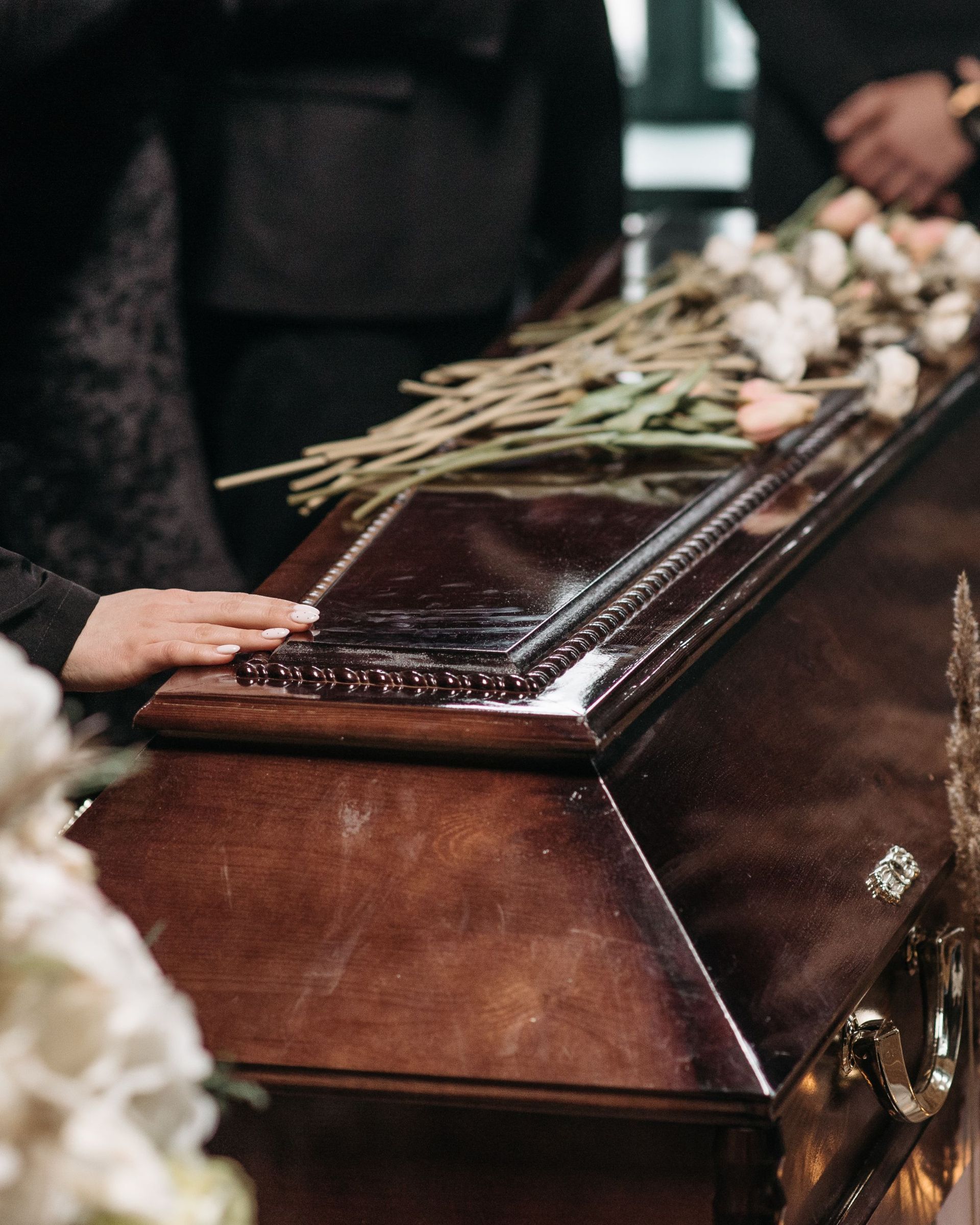 Close-up of a brown casket with flowers on top; a hand rests on the lid.