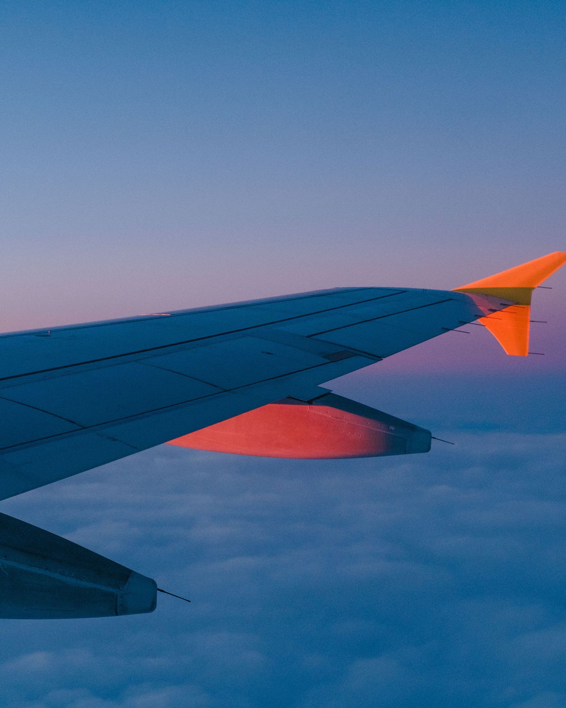 Airplane wing against a sunset sky over clouds, orange and purple hues.