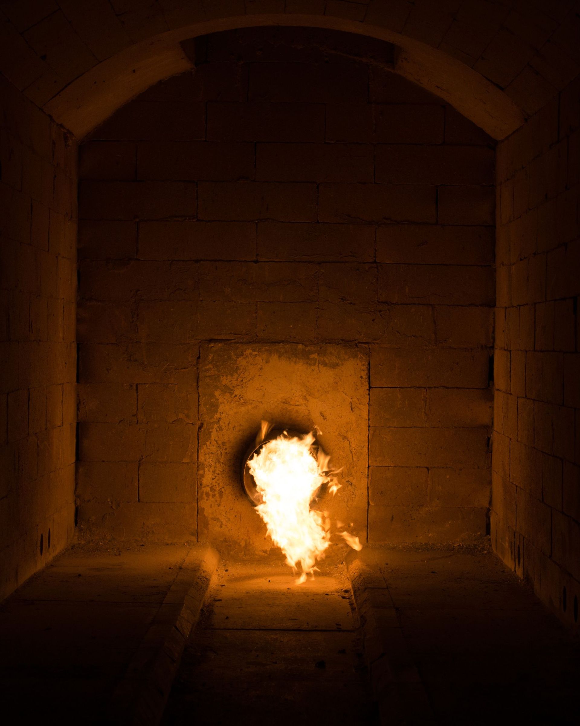 Inside of an oven, a bright flame erupts from a circular opening, casting light on the brick walls.