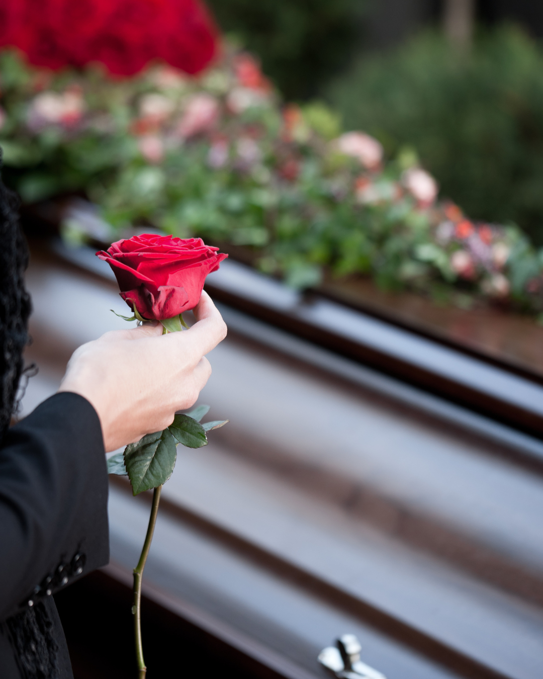 Person's hand holding a red rose, placing it on a closed brown casket decorated with flowers.
