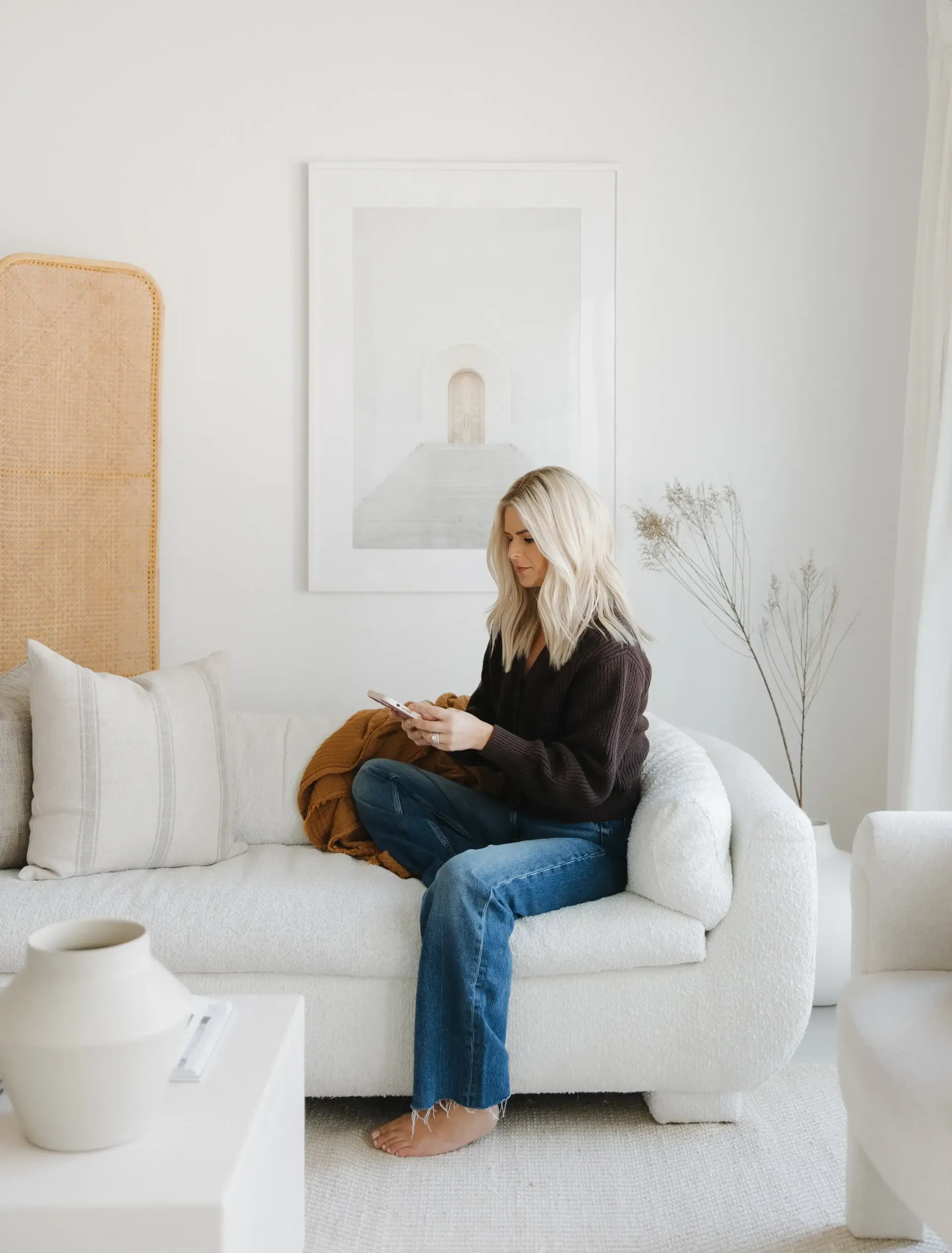 A woman is sitting on a white couch in a living room looking at her phone.