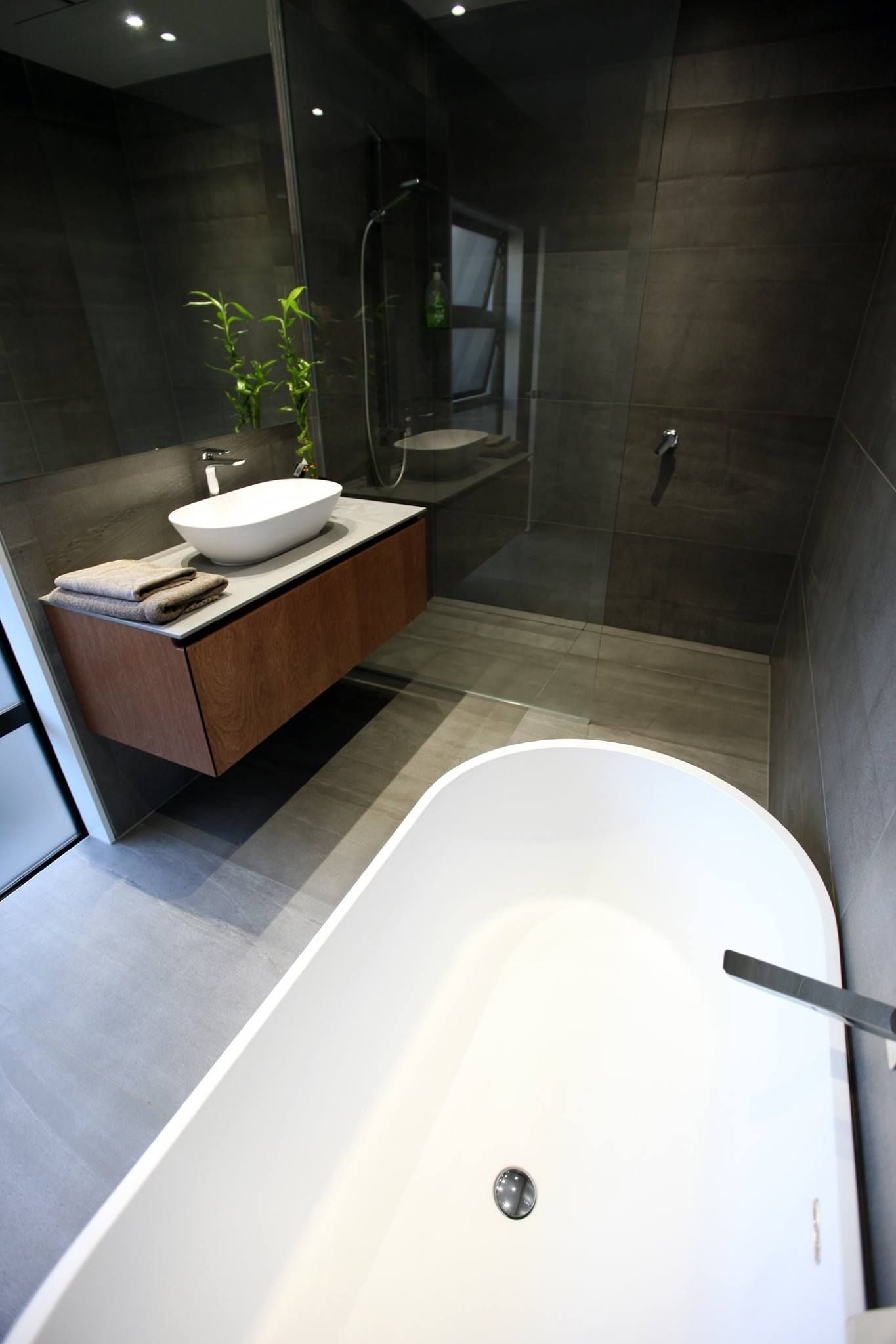 A modern, minimalist bathroom with a white freestanding bathtub in the foreground and a floating wooden vanity behind it.