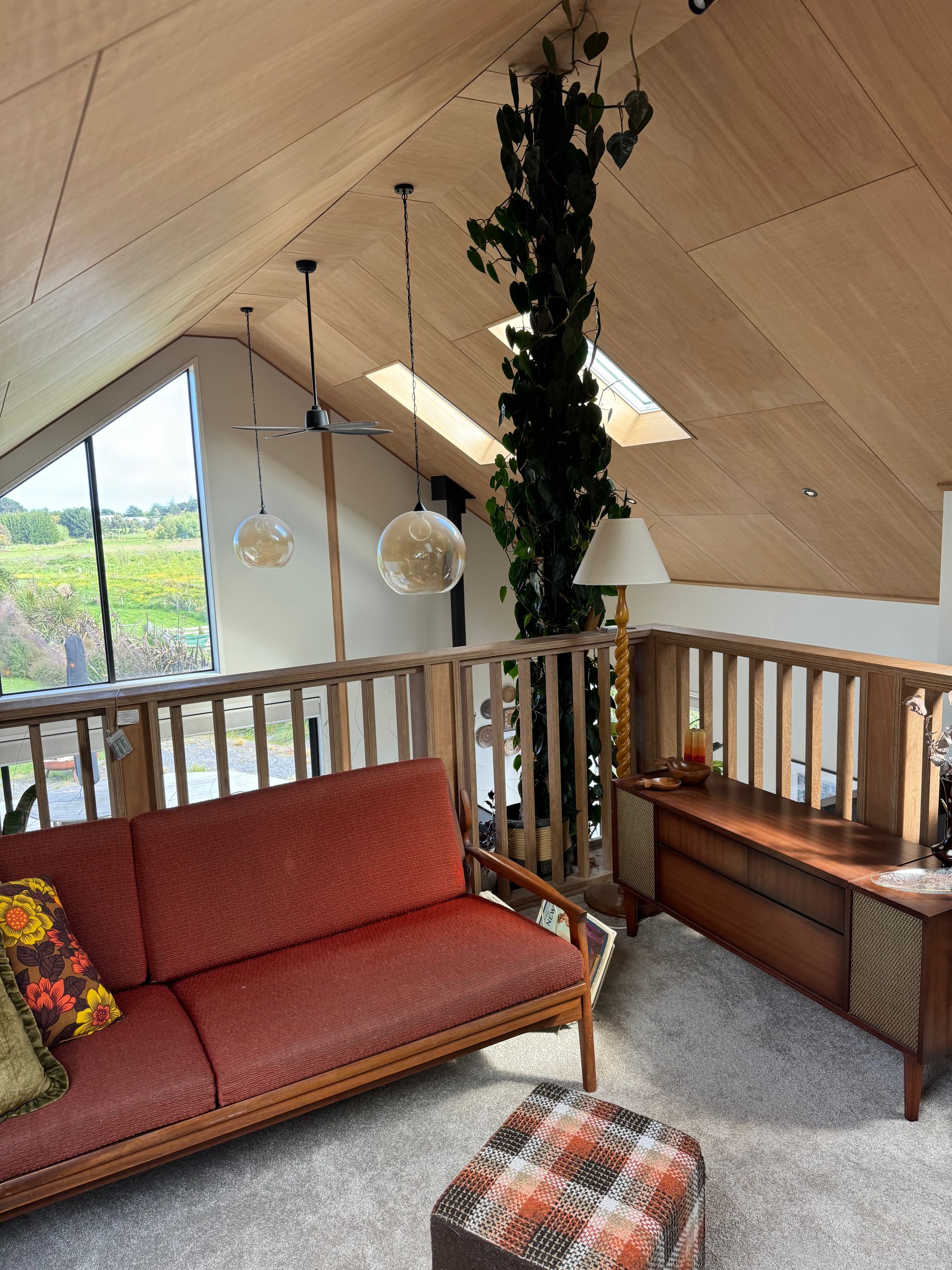 An attic living area with a red sofa, a wooden sideboard, and a tall, leafy indoor plant under a sloped wooden ceiling.