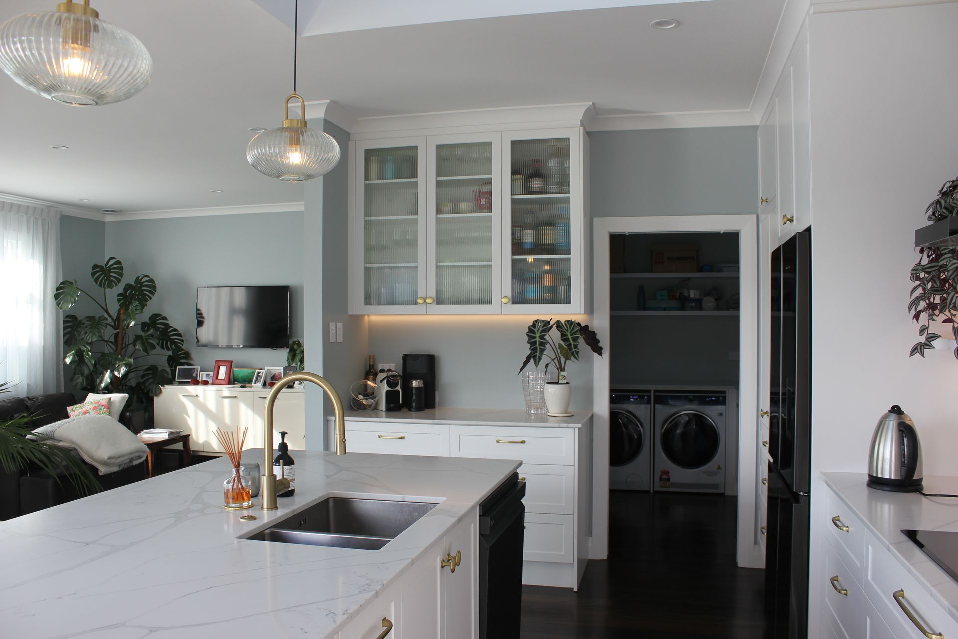 A modern white kitchen with a marble-topped island, hanging lights, glass-front cabinets, and an adjacent laundry room.
