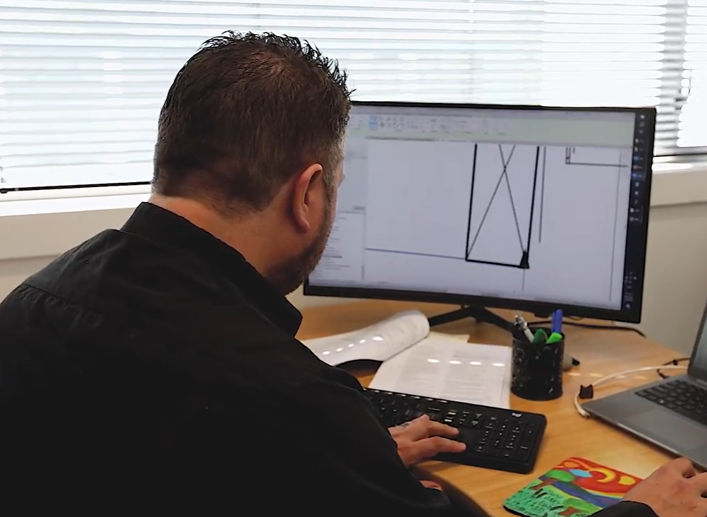 A person sits at a desk working on a computer screen showing a technical design, with a keyboard and papers nearby.