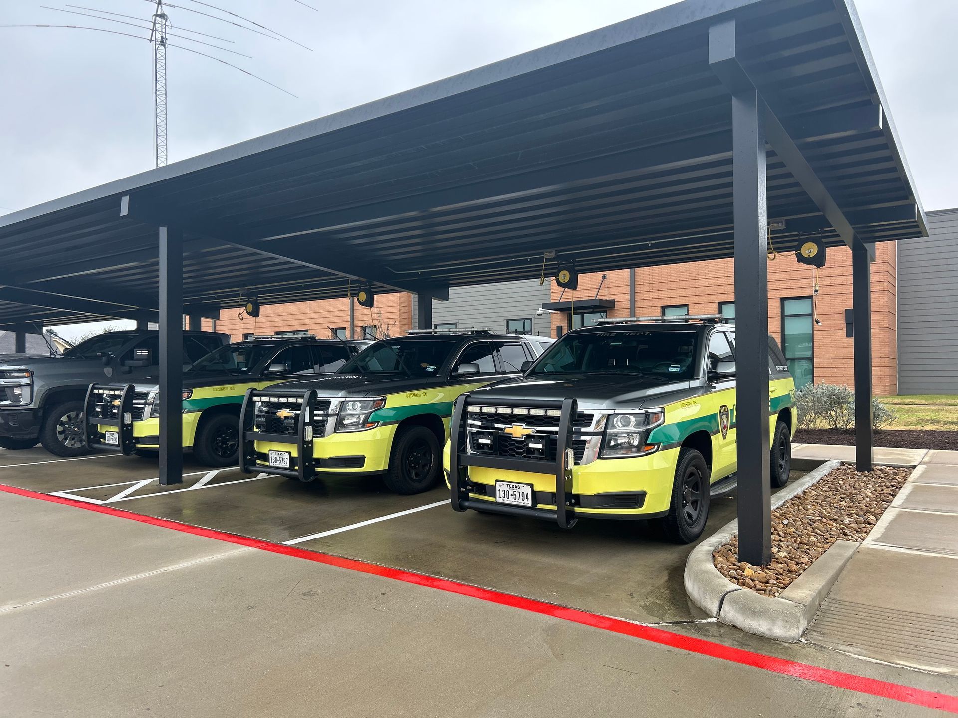Three light-green and black patrol SUVs parked in a row under a metal carport outside a brick building.