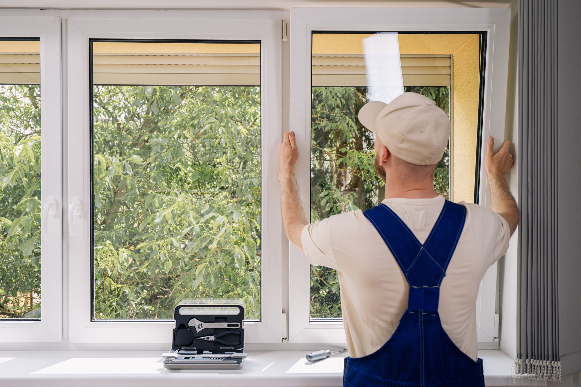 A man in overalls installs a window; tools on windowsill, trees outside.