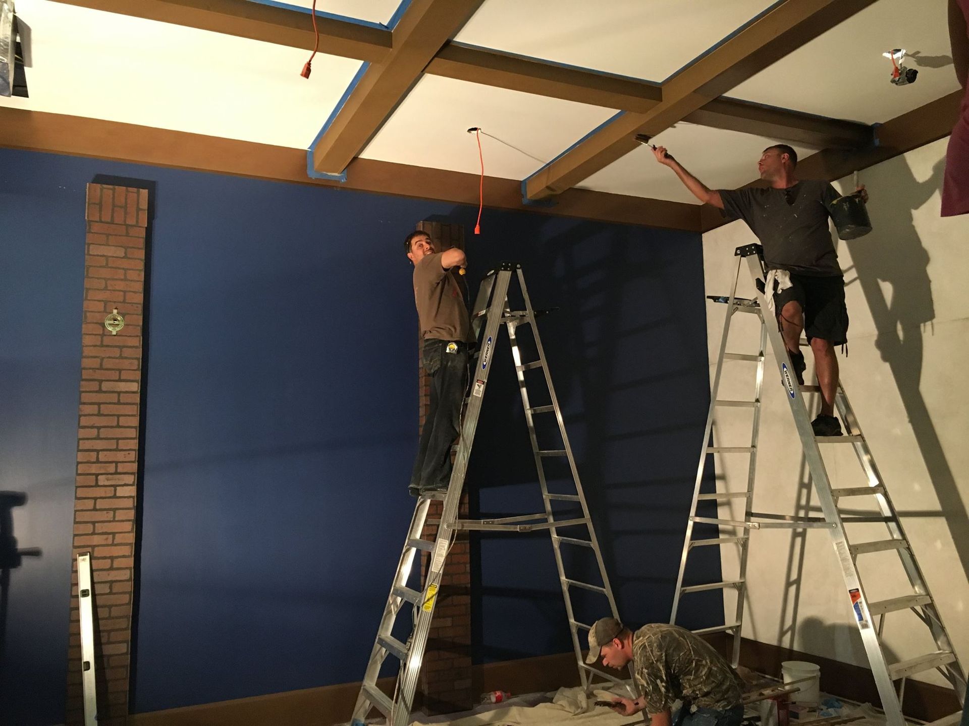 Three people painting a room, one on a ladder touching a beam, another on a ladder, and one on the floor.