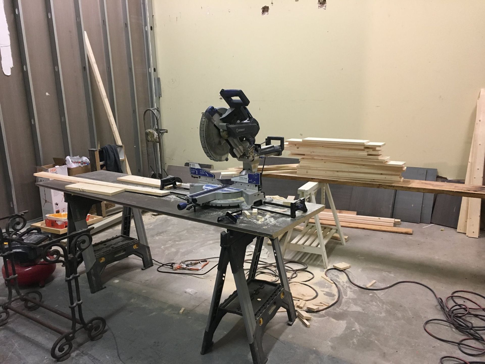 Miter saw on a workbench, surrounded by lumber, air compressor, and other tools in a workshop.