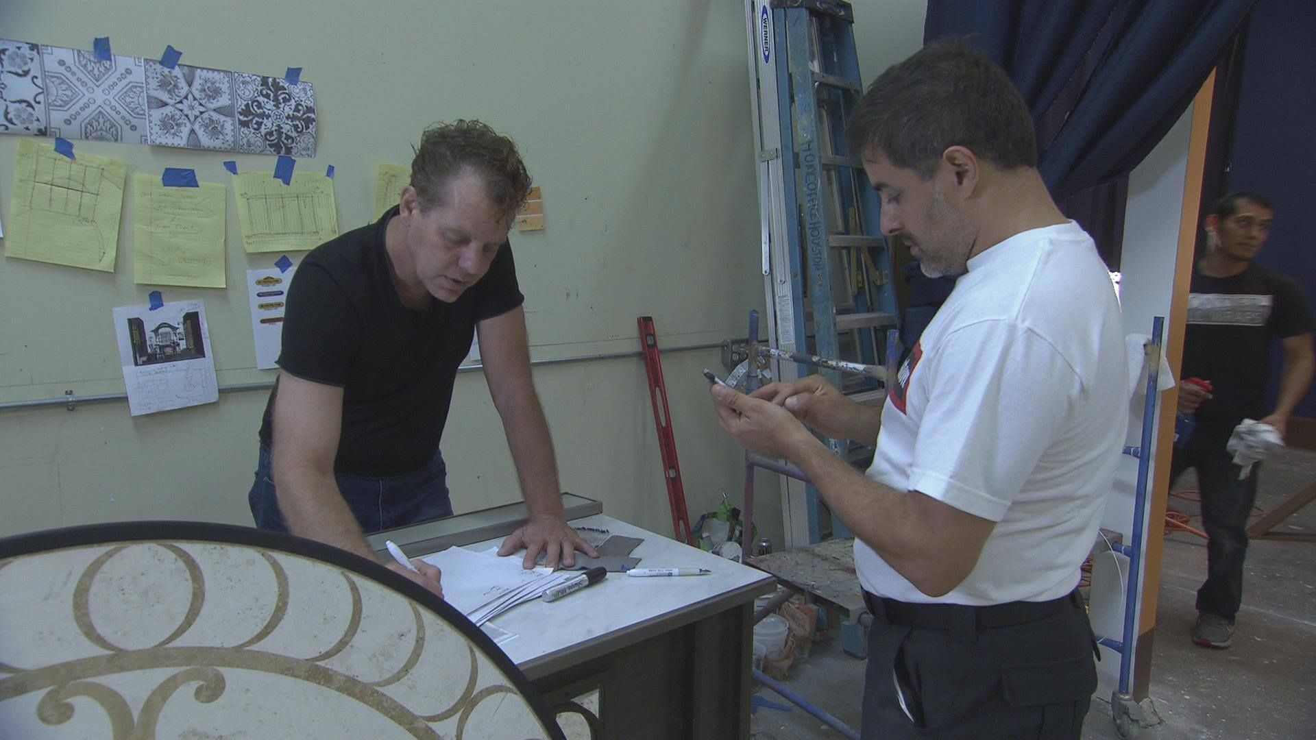 Three men working on a project; one cuts tiles, another examines them, and a third carries something.