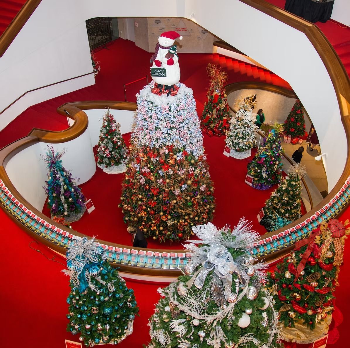 A circular hall filled with decorated Christmas trees, viewed from above, with red carpet and a central tree adorned with a snowman.