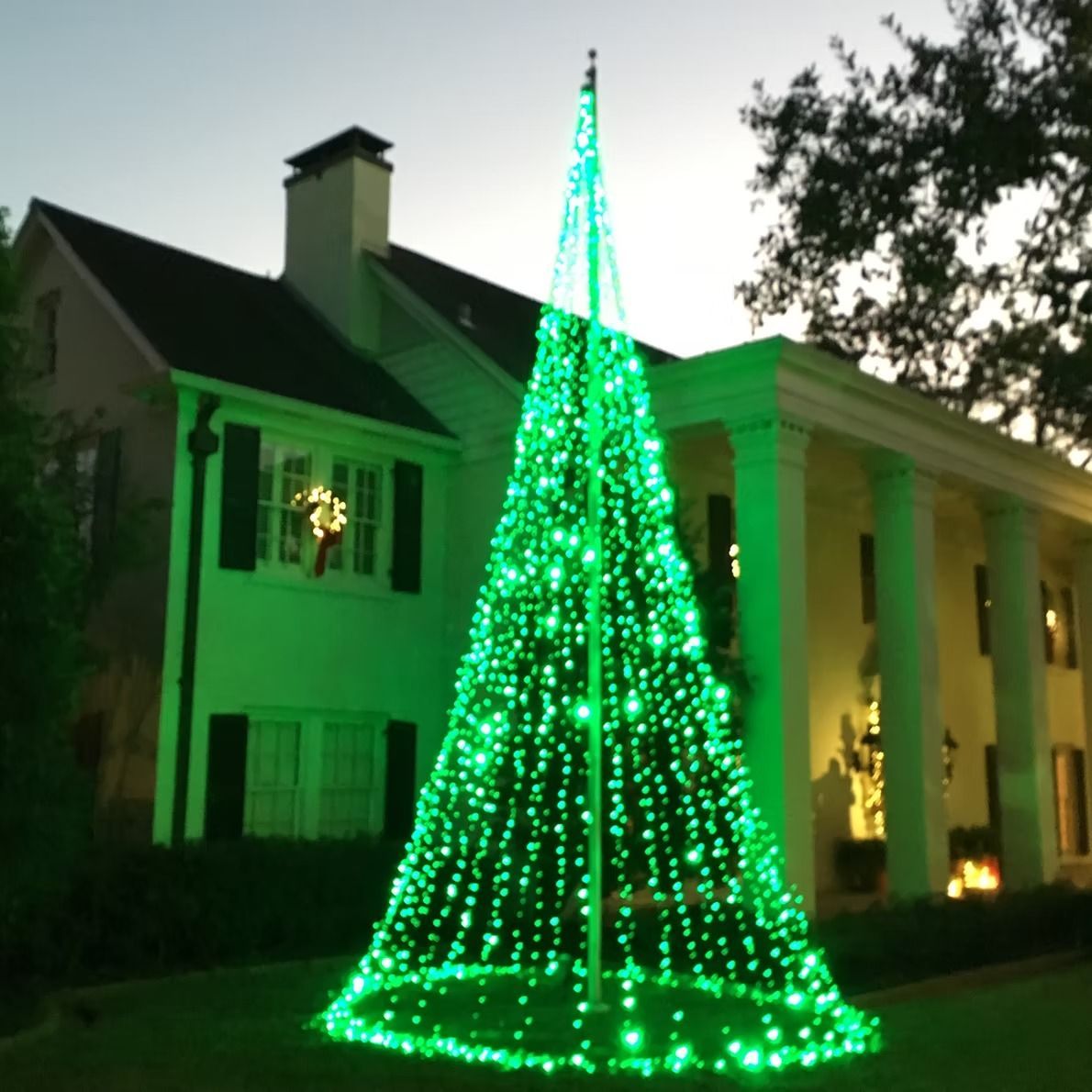 Large, glowing green Christmas tree made of lights in front of a white house with columns.