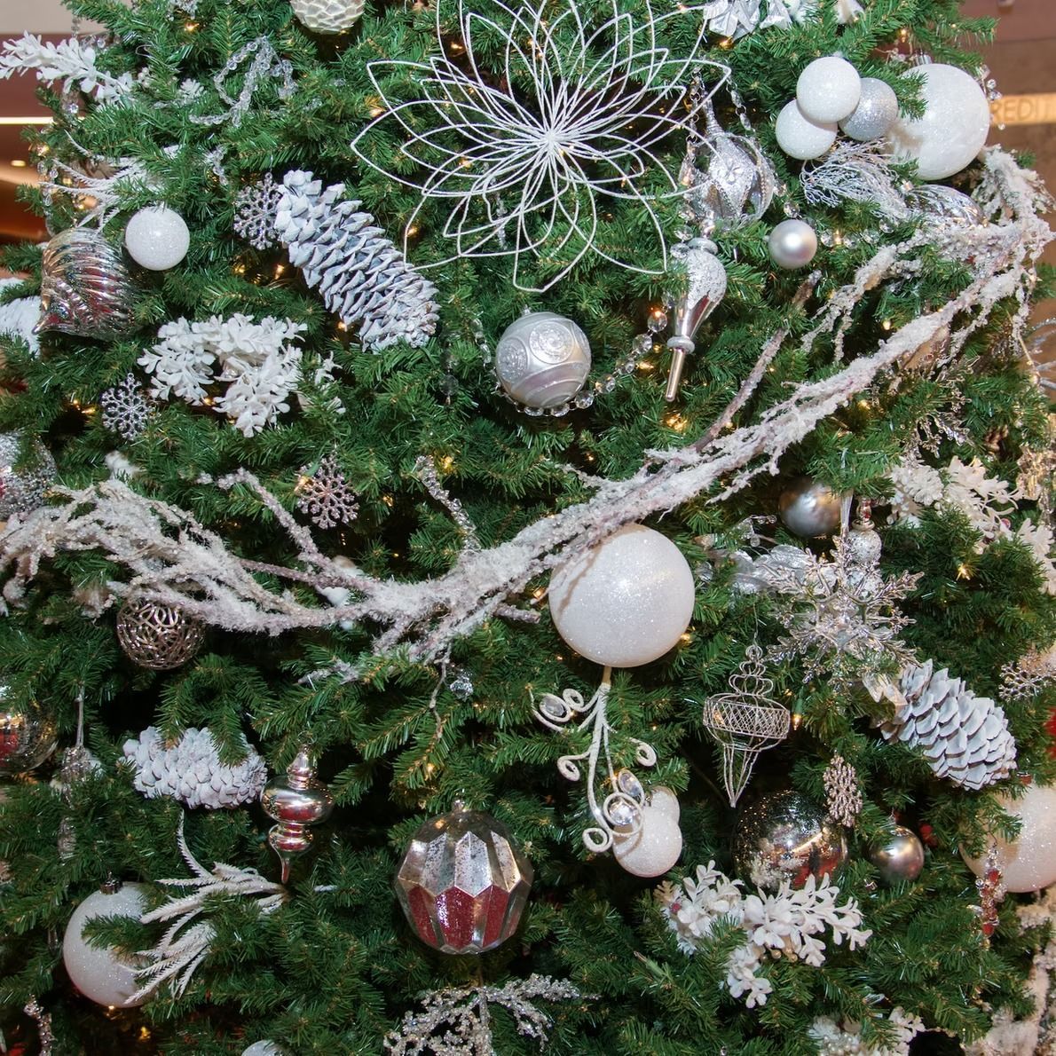 Close-up of a decorated Christmas tree with silver and white ornaments, faux snow, and a large silver floral accent.