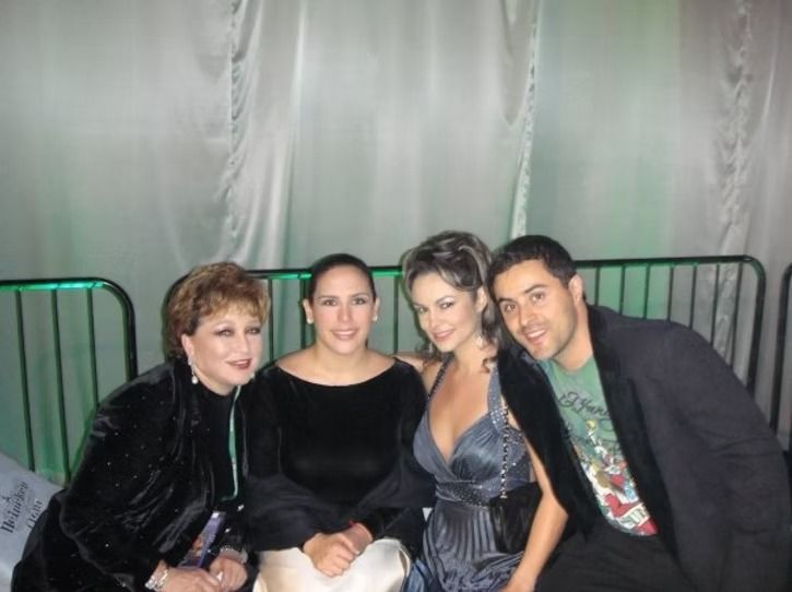 Four people posing, smiling. The woman in blue dress has an arm around the woman next to her. Metal fence background.