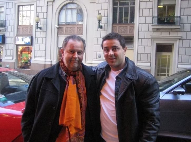 Two men standing outside a building, one wearing a black coat and orange scarf, the other in a leather jacket.