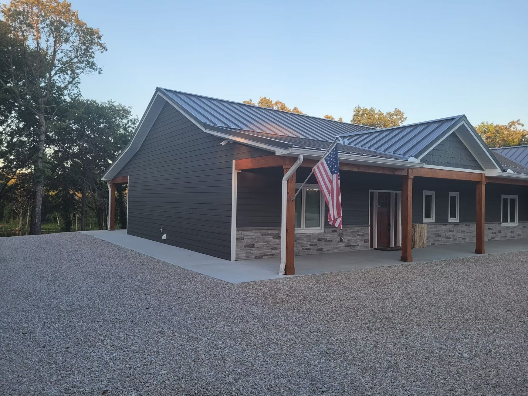A modern one-house with a roof, dark siding, and a covered porch with an American flag. The house sits on a gravel driveway.