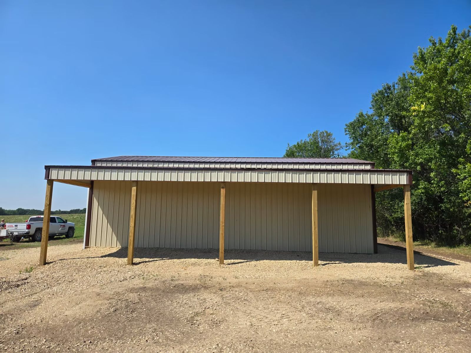 A beige metal shed with a brown roof and a covered porch, set on a gravel lot under a blue sky.