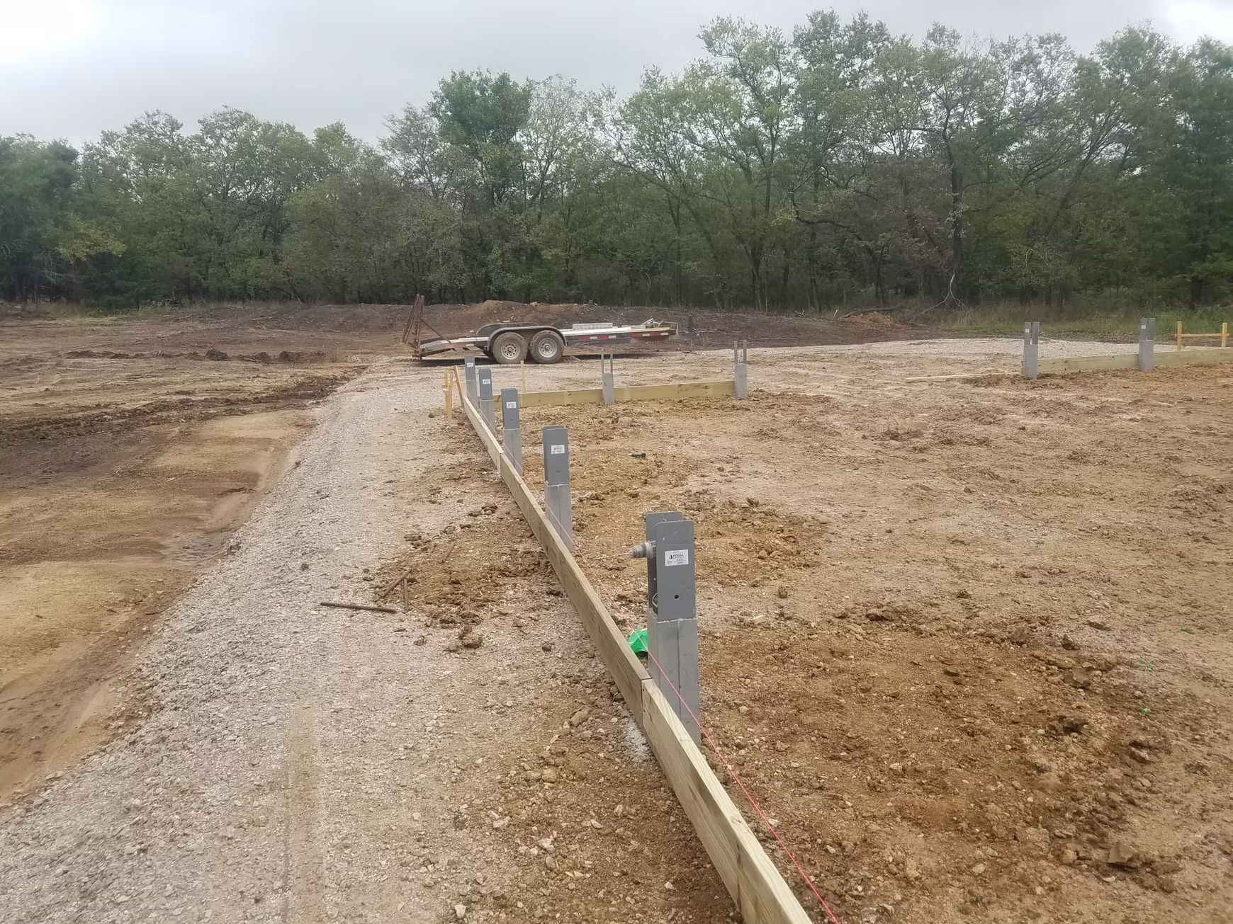 Construction site with a wooden beam and metal posts, gravel border, and dirt. Trees in the background, trailer visible.