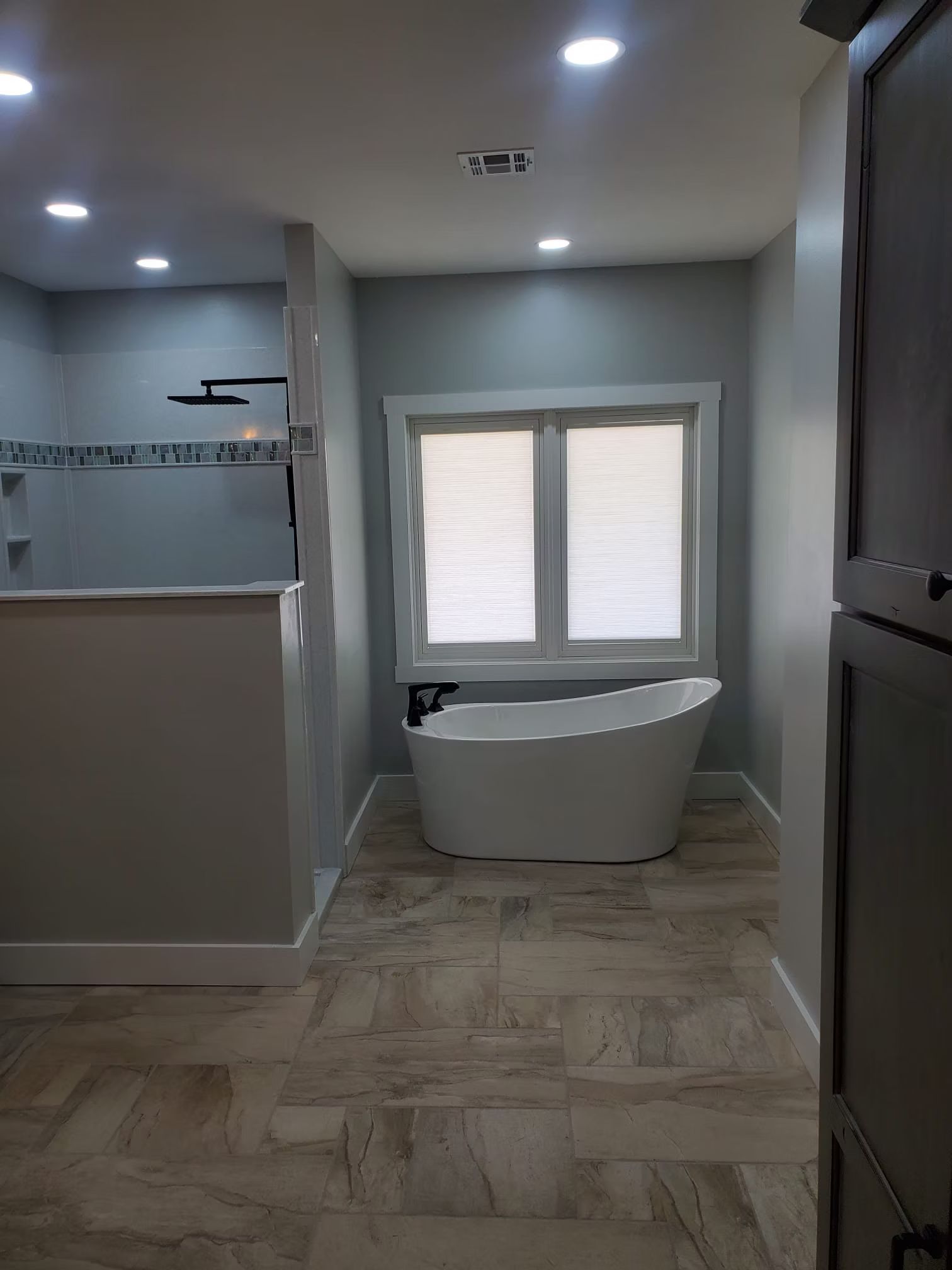 A modern bathroom with a freestanding bathtub under a window. Light gray walls and flooring, with a walk-in shower visible.