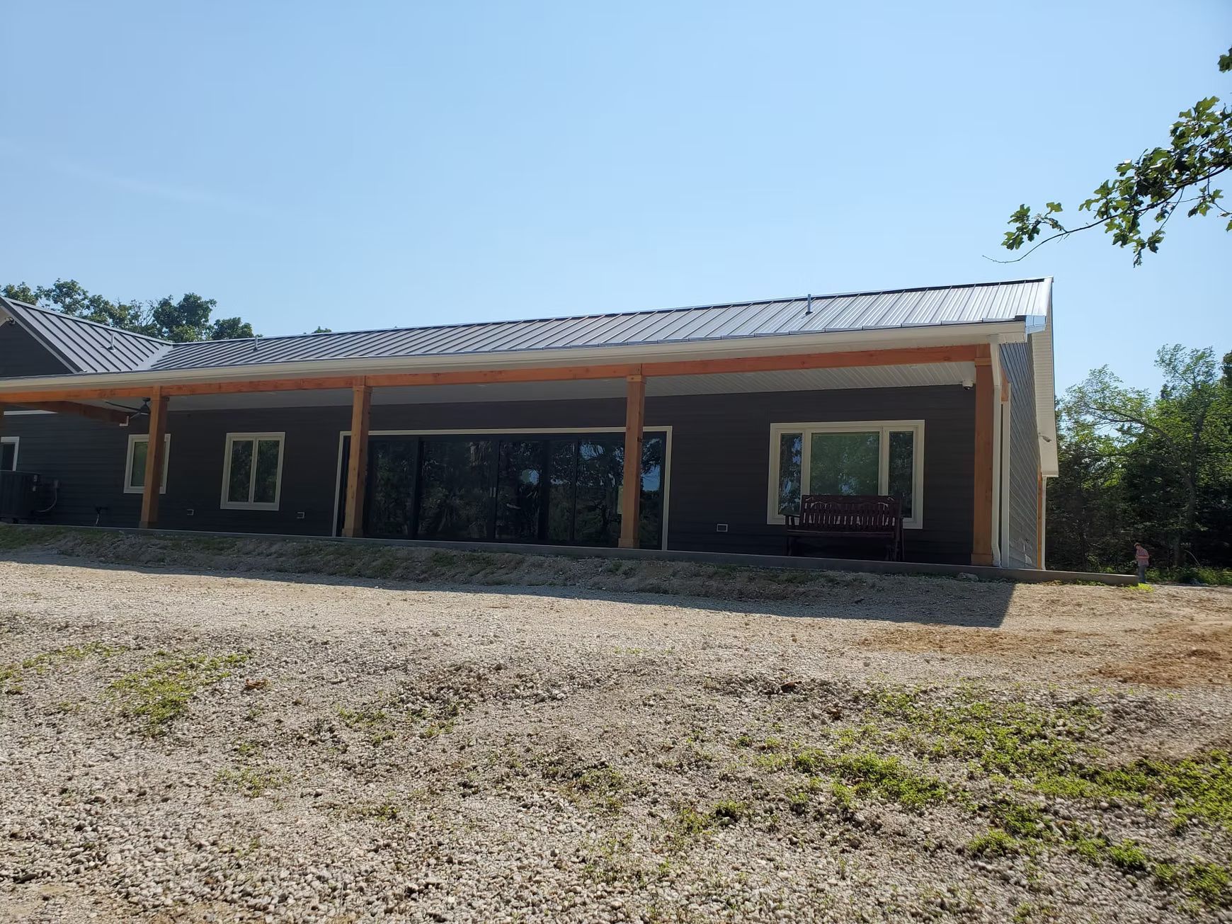 A one-story building with a dark gray exterior and a covered porch supported by wooden beams, with a gravel yard i\