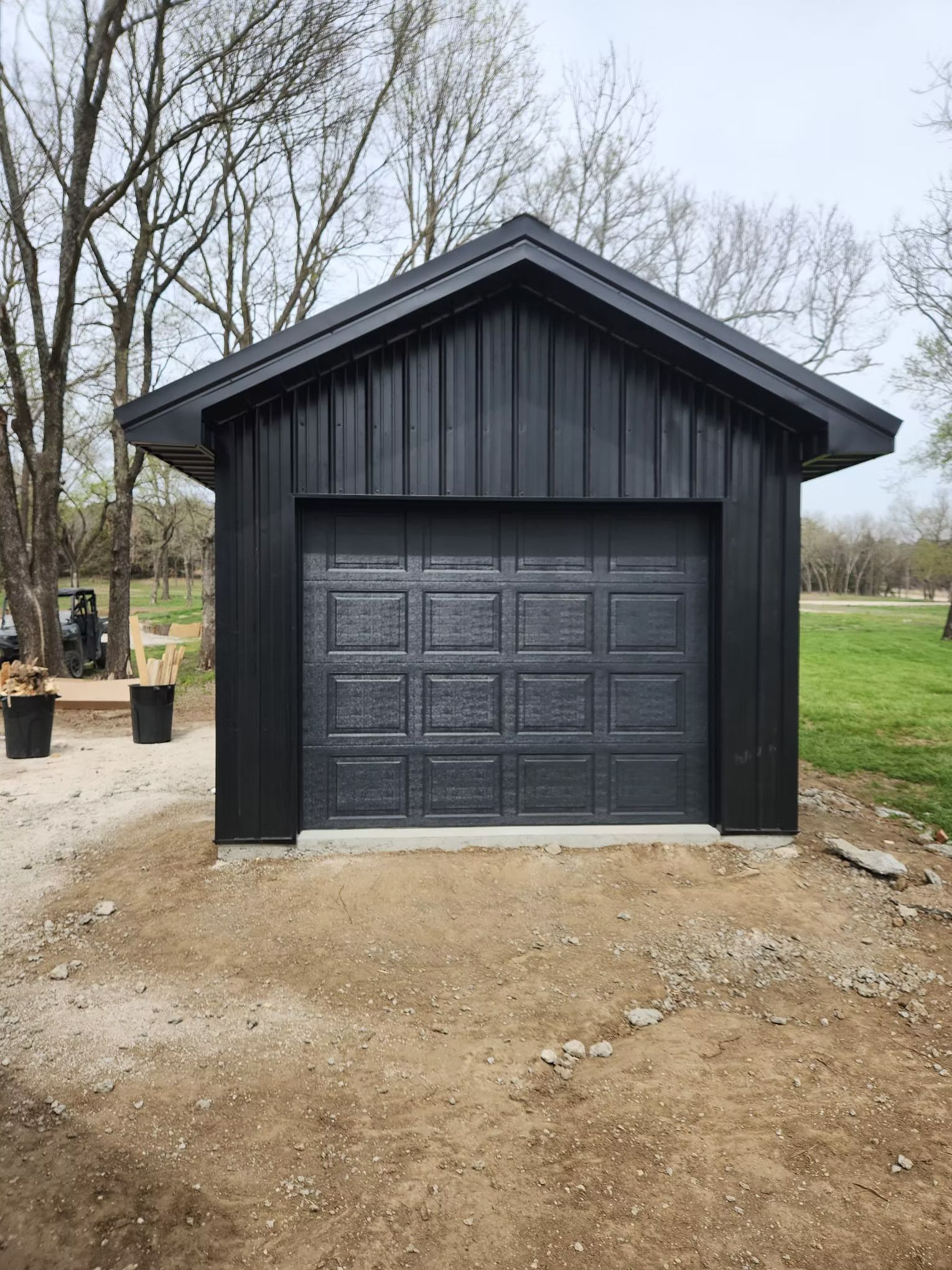 Black metal garage with a matching door and roof against a backdrop of trees and a dirt lot.