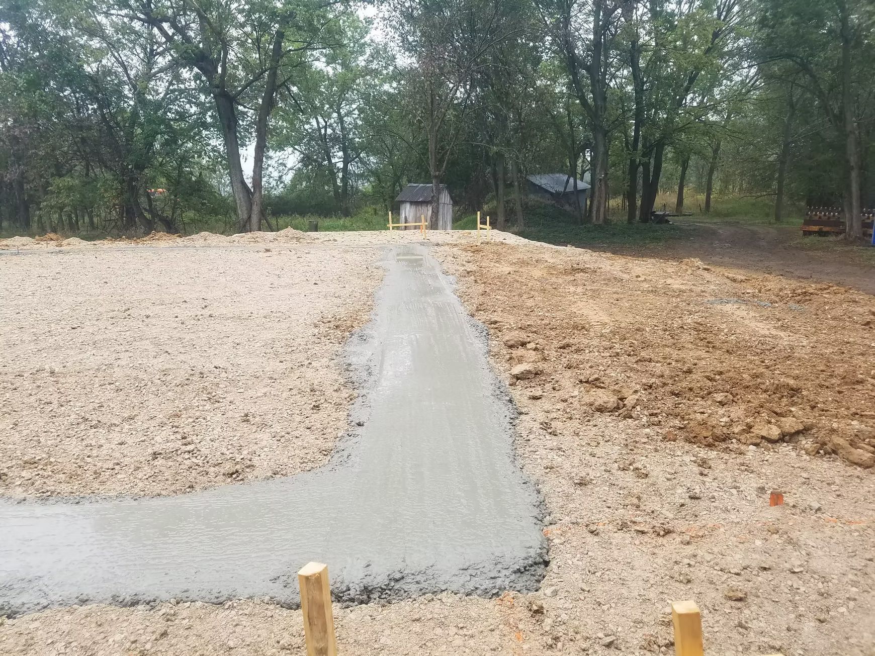 A freshly poured concrete path snakes through a gravel-covered construction site, leading toward trees in the background.