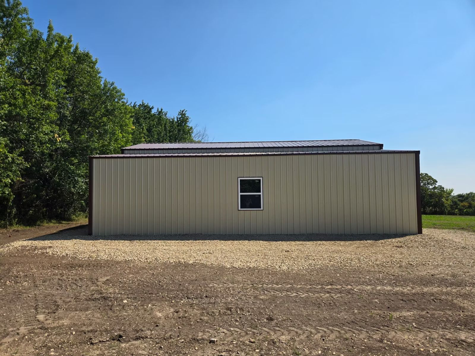 Beige metal shed with brown trim and a small window, set against a blue sky and gravel ground.