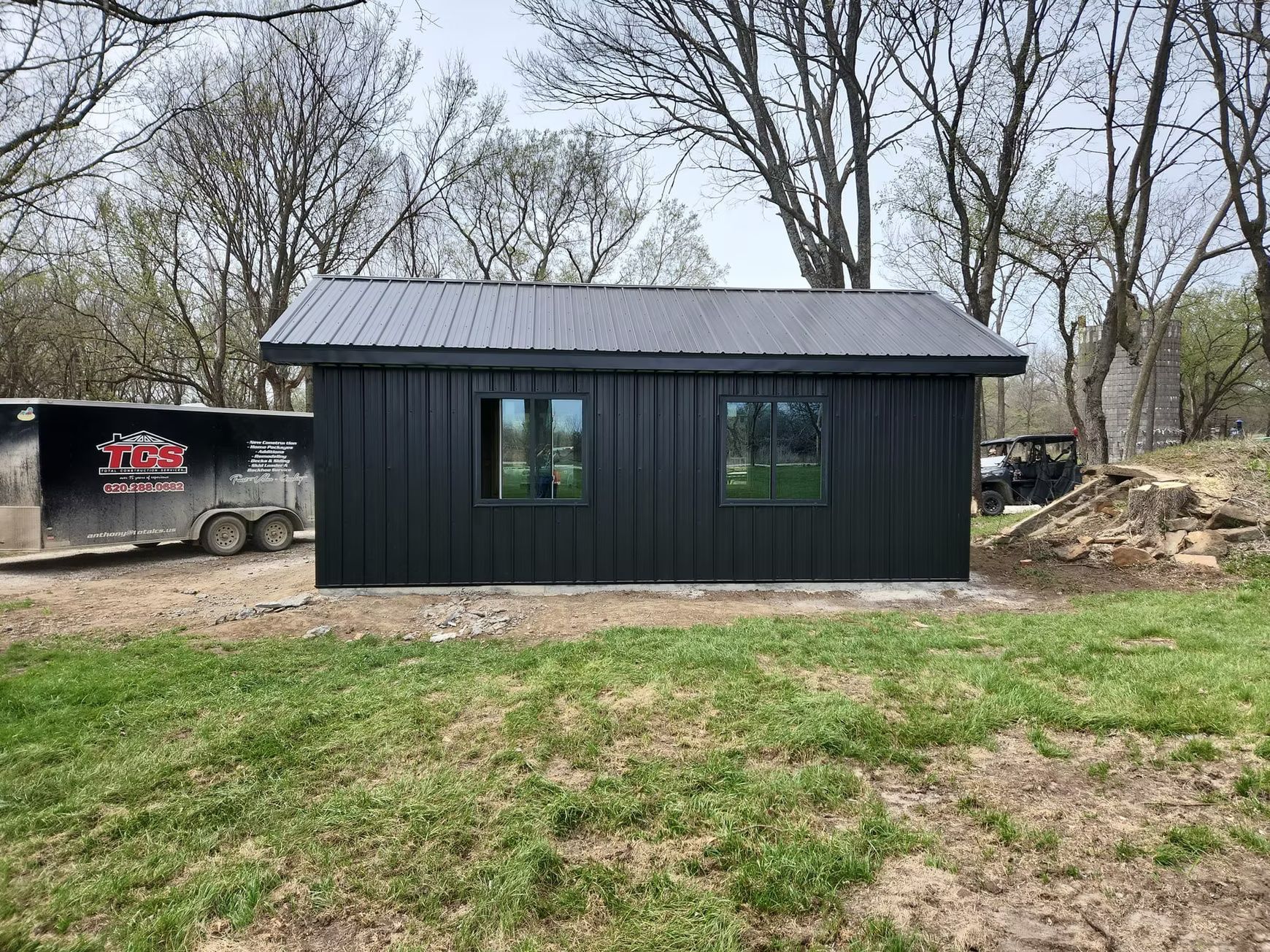 Black shed with a metal roof and two windows, set in a grassy yard with trees in the background. A trailer sits to the left.