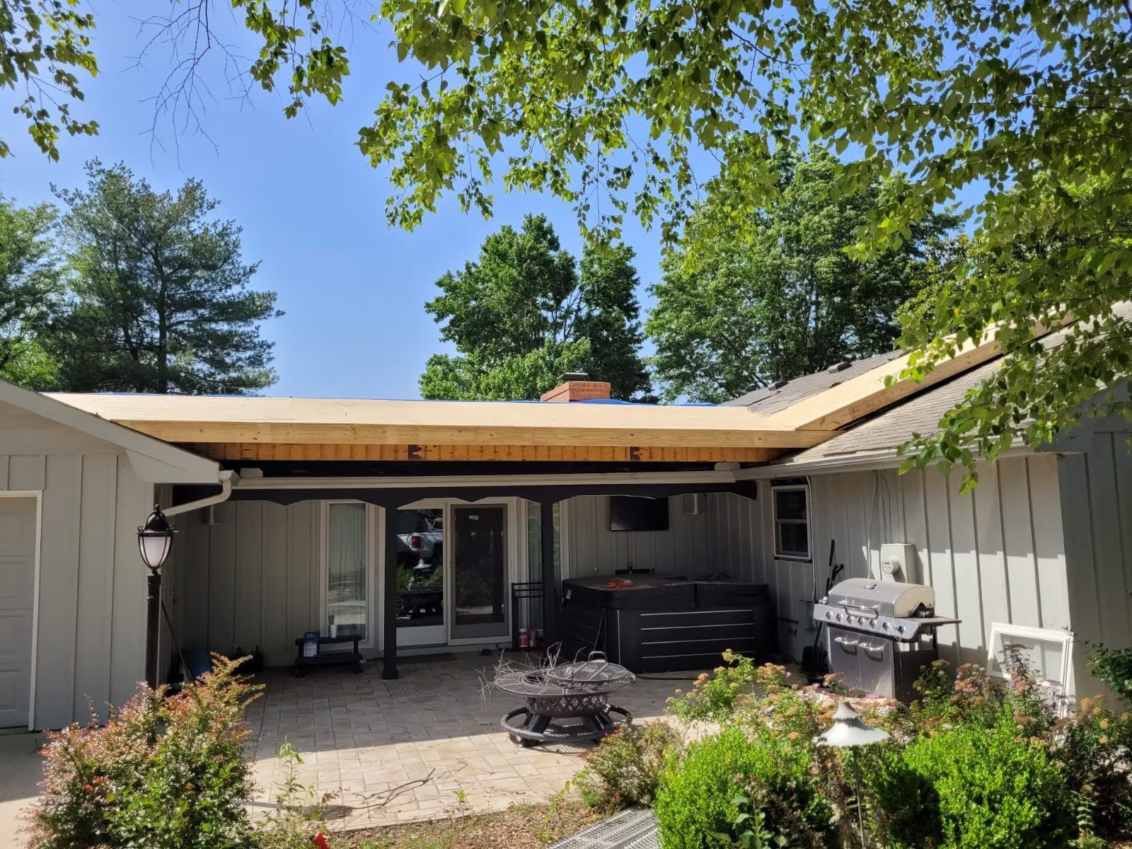 Backyard with a flat A hot tub, grill, and patio furniture are visible. Green trees and a blue sky fill the background.