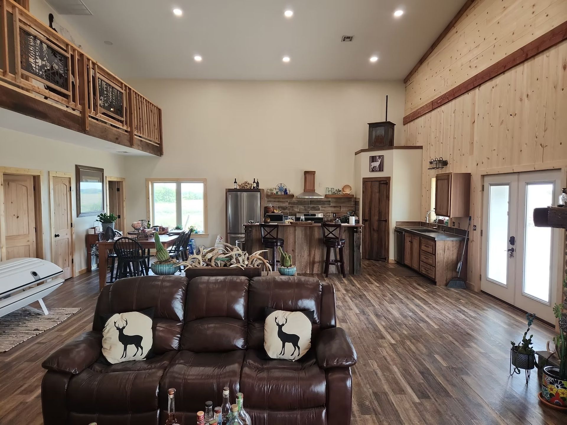 Open-concept living space with a leather sofa, wood flooring, and a kitchen area. The walls are light-colored wood.
