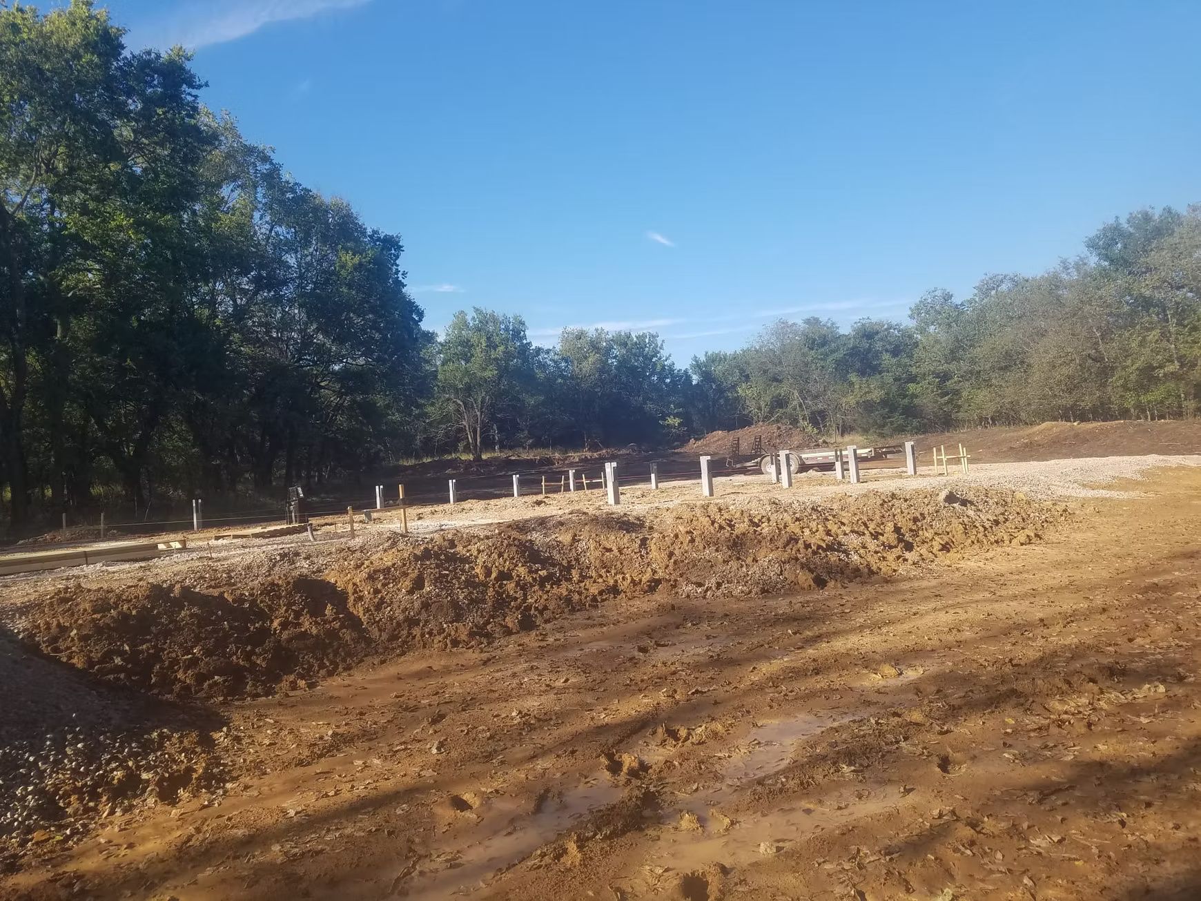 Construction site with exposed concrete pillars and earth mounds, surrounded by trees under a blue sky.