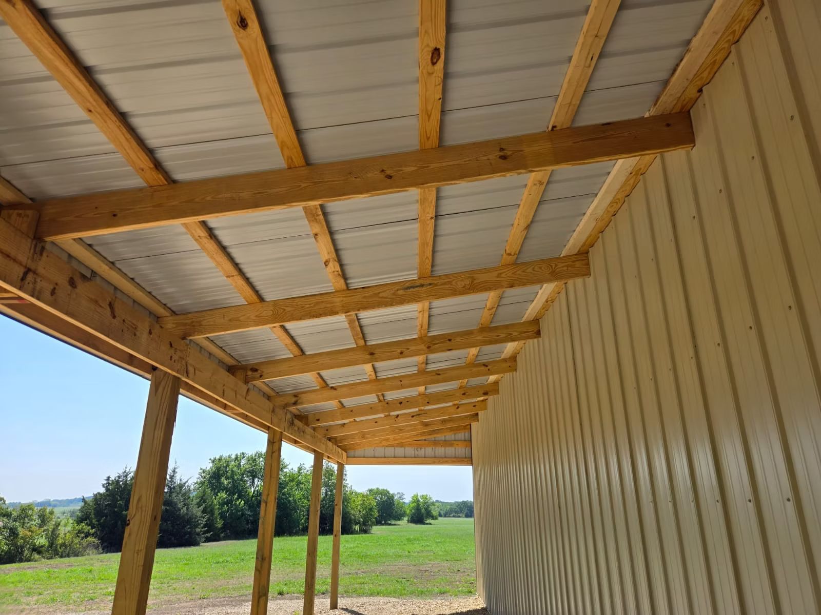A wooden porch with a metal roof attached to a beige building. Sunlight illuminates a grassy field in the distance.