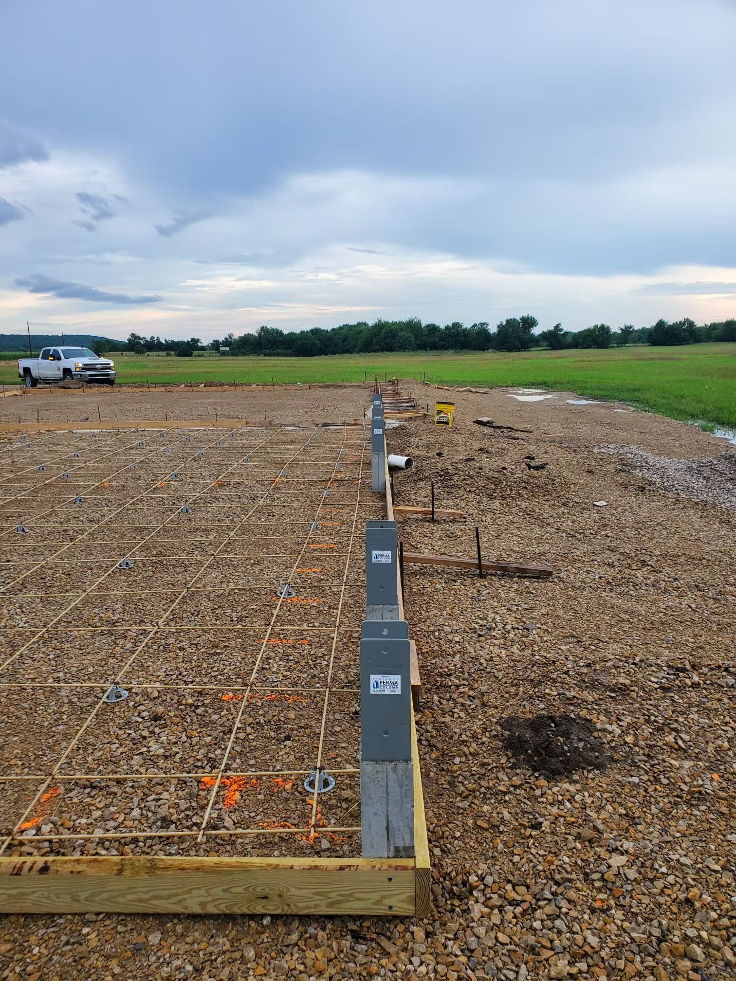 Construction site with gravel base, wooden forms, and reb truck is visible in the distance under an overcast sky.