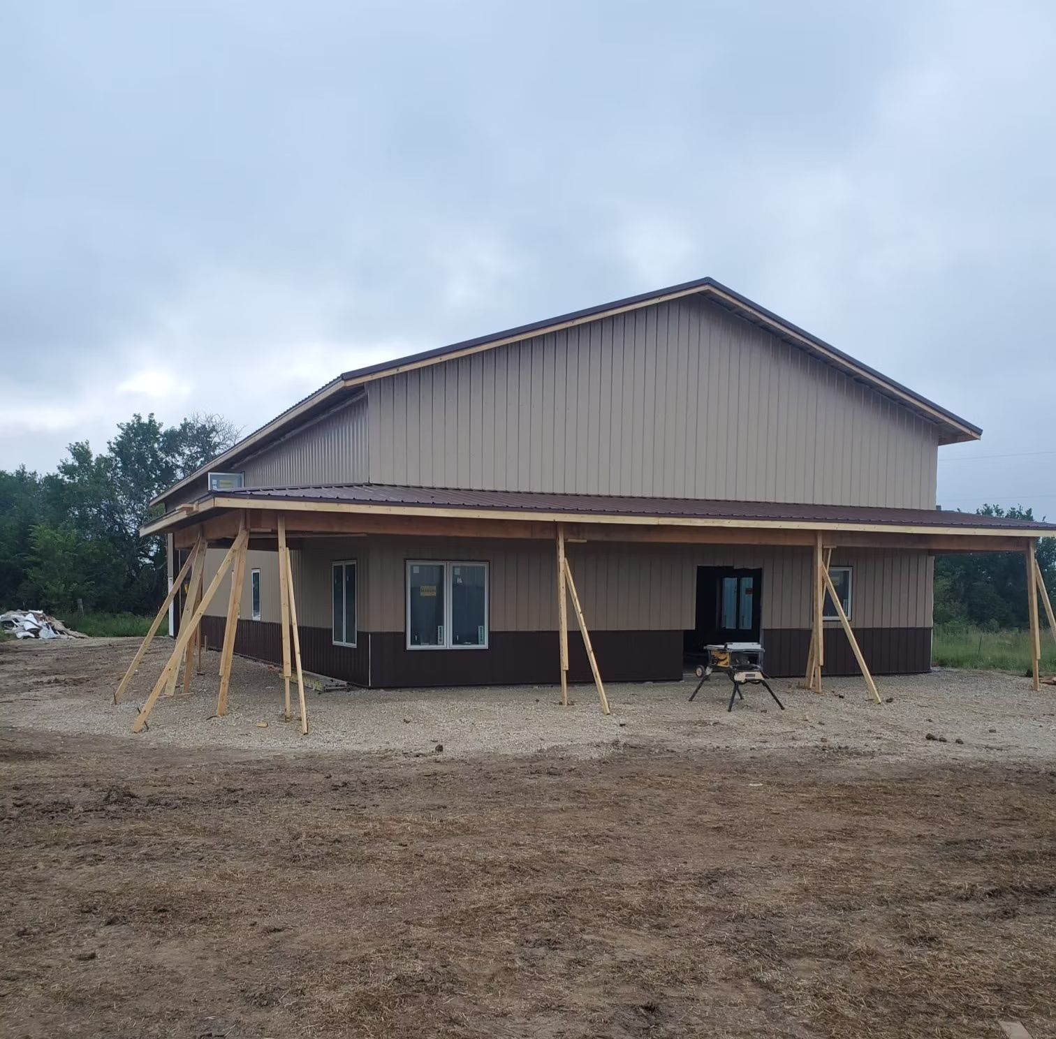 A two-story building under construction, with tan and brown a covered porch supported by wooden posts, set on a gravel lot.
