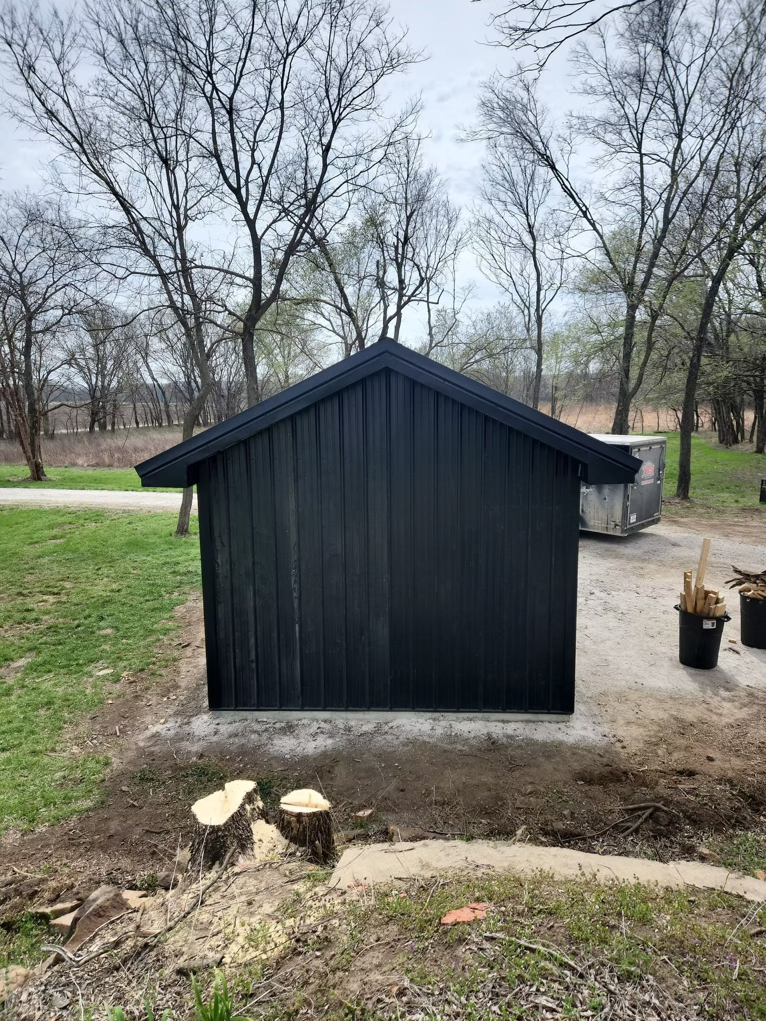 A dark-colored, small building with a gabled roof sits on a concrete pad. Trees are in the background.