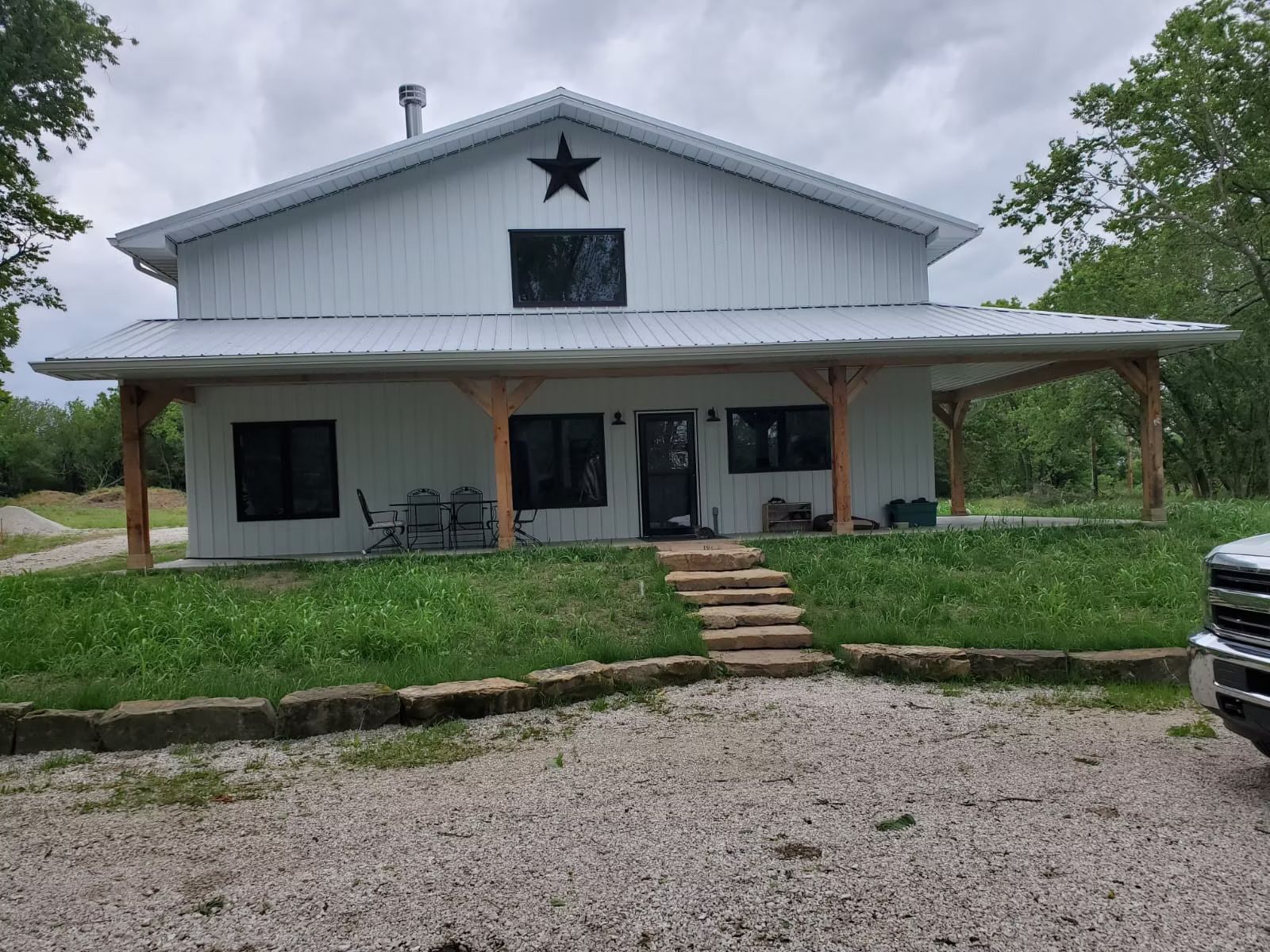 White, two-story be house with a star decoration, porch, and gravel driveway. Green grass and trees surround the home.