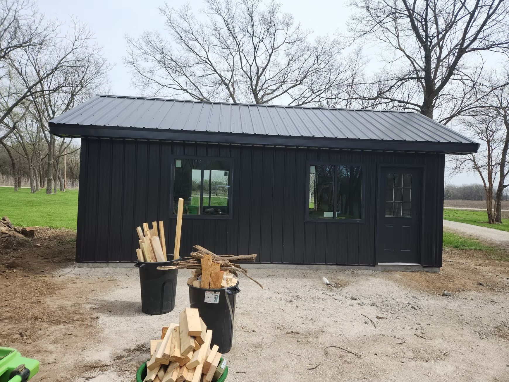 A black shed with a metal roof sits on a gravel lot, surrounded by trees.a door are visible. Firewood rests in containers.