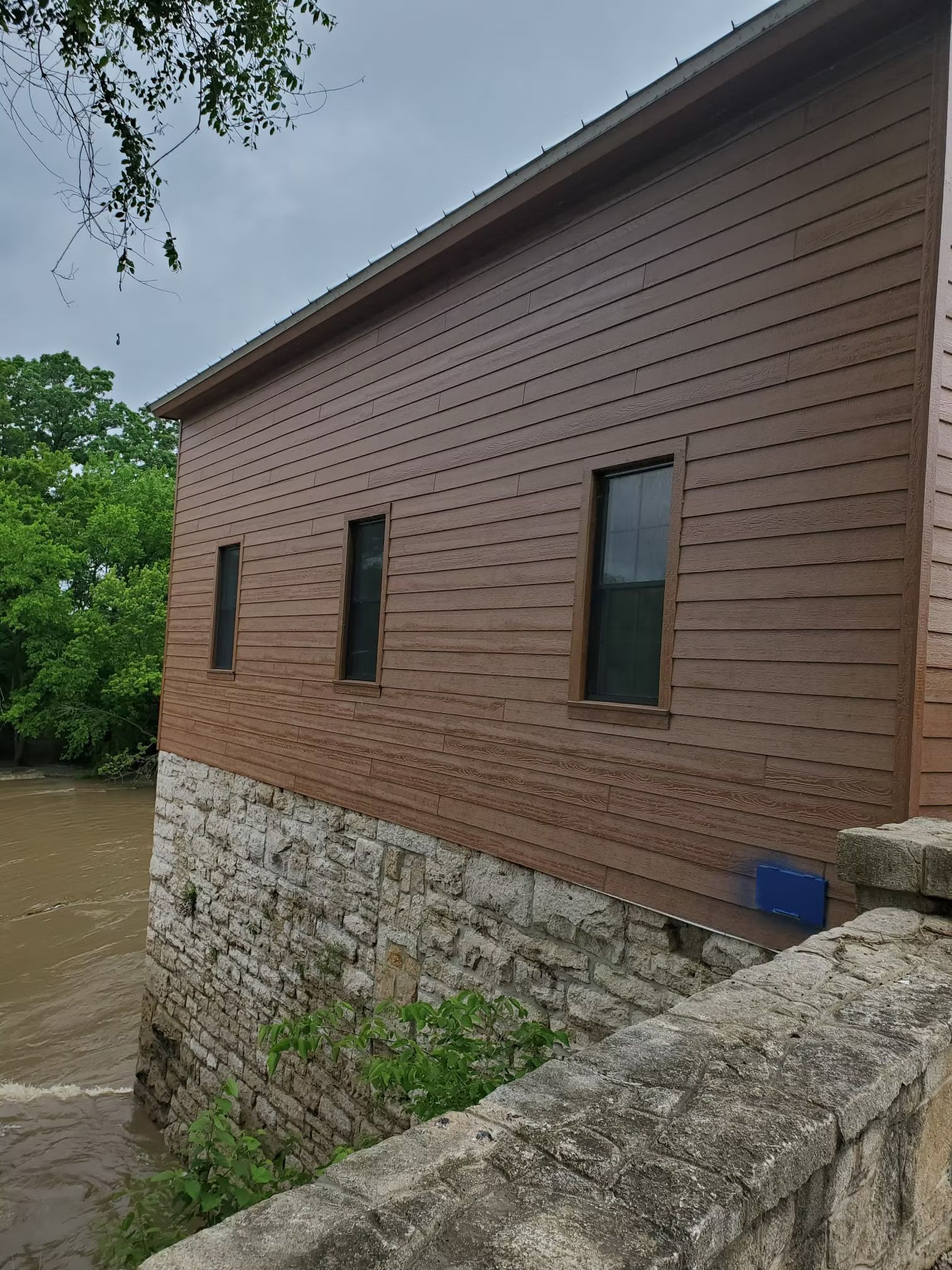 Brown wooden-sided building with three windows sits atop a stone foundation. Water is visible below the building.