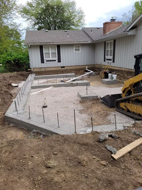 Construction site with a cono a light gray house.  Rebar sticks up from the concrete. A small excavator is visible.