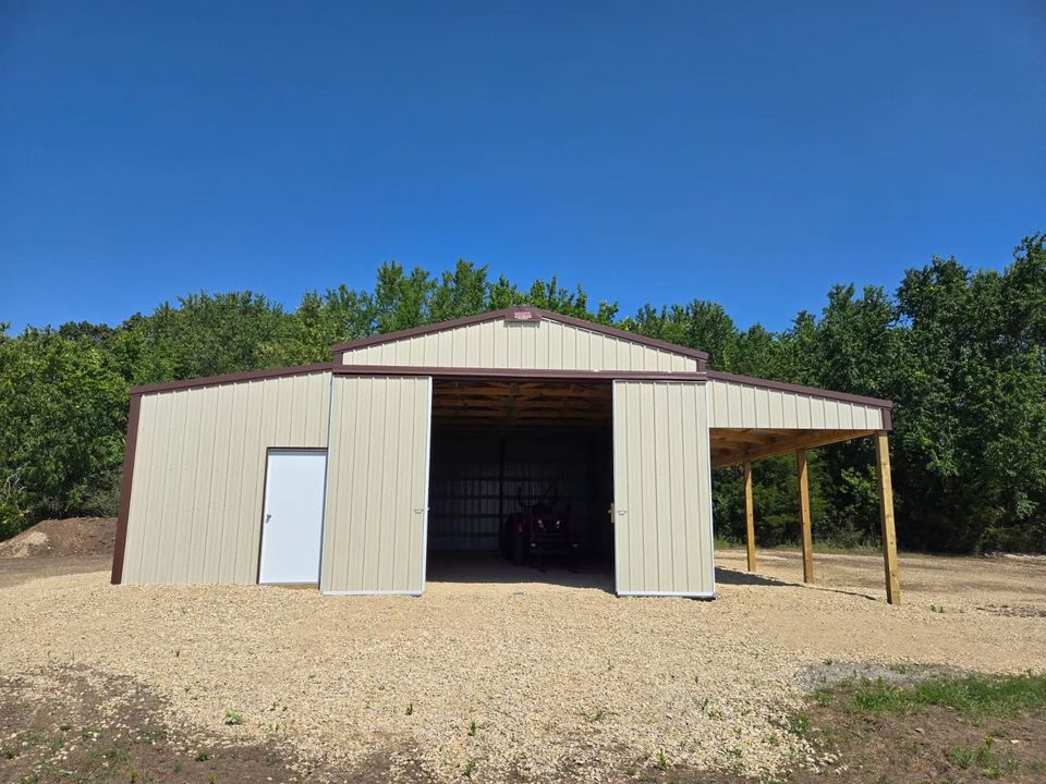 Tan metal barn with brown trim and an open front entrance, a side overhang, and gravel ground.