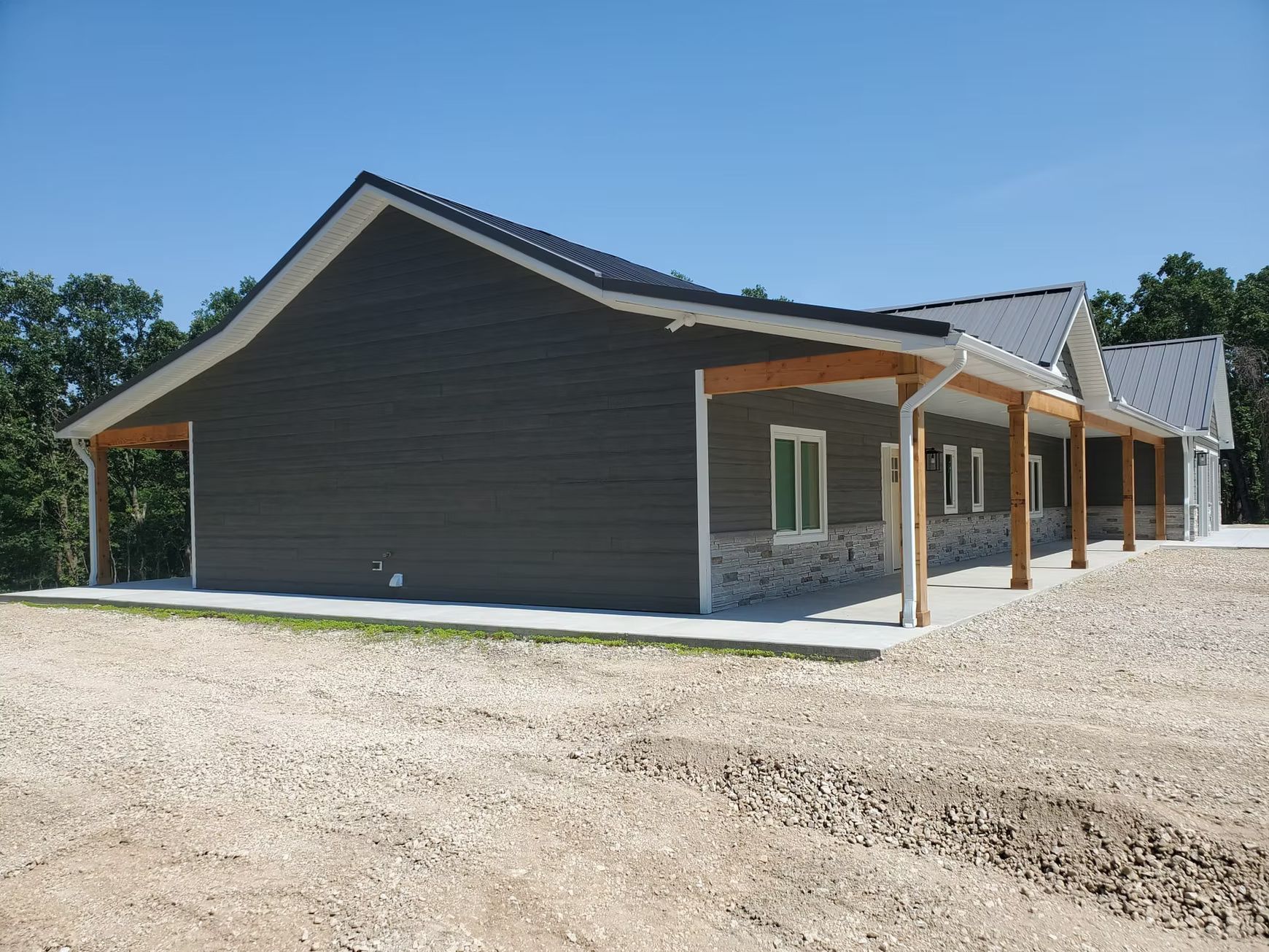 New building with dark gray siding, a porch, and a gravel lot. The roof is black and the sky is blue.