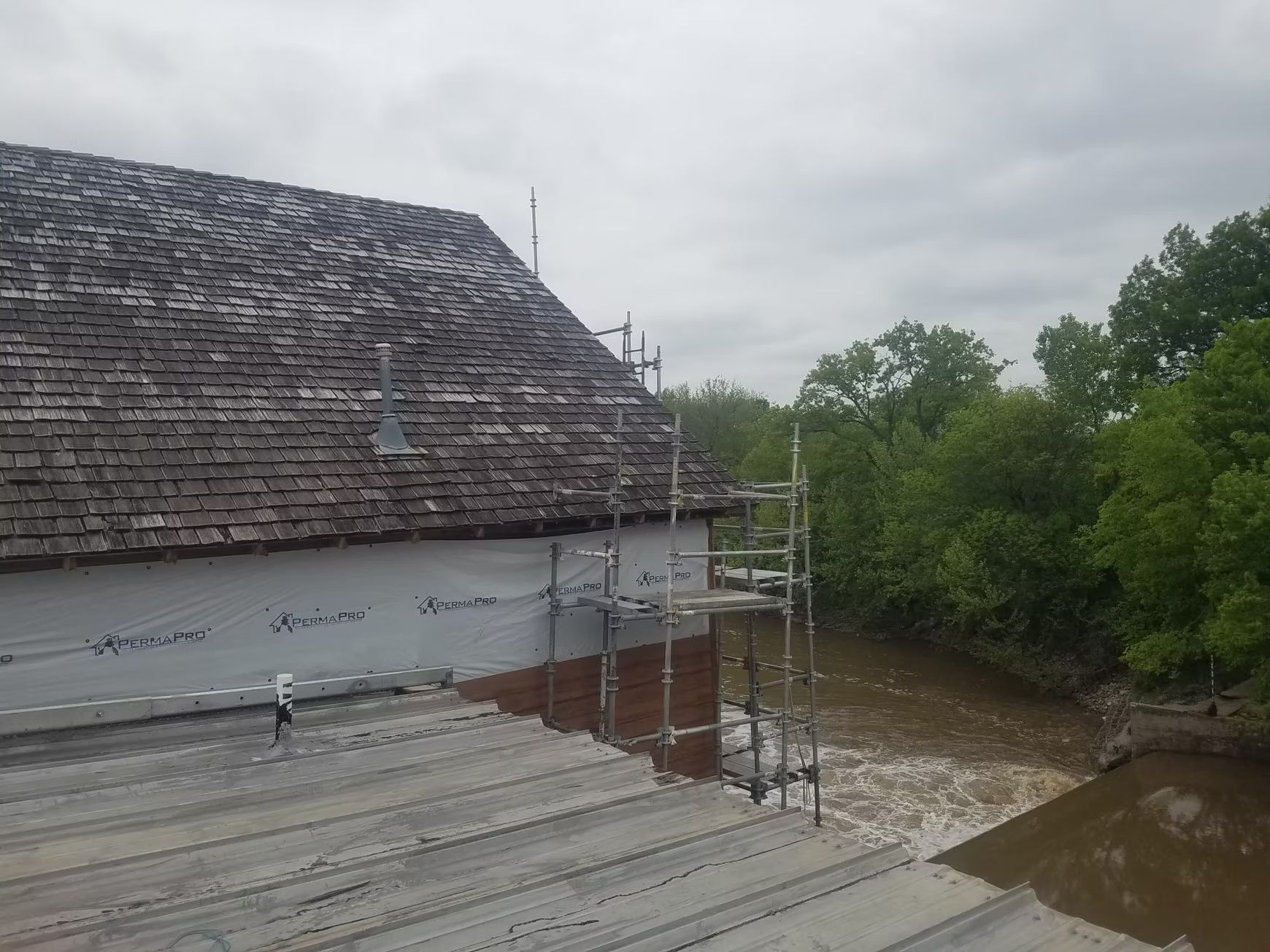 A building with a weathered roof sits near a flooded area with trees. Scaffolding lines the side of the building.