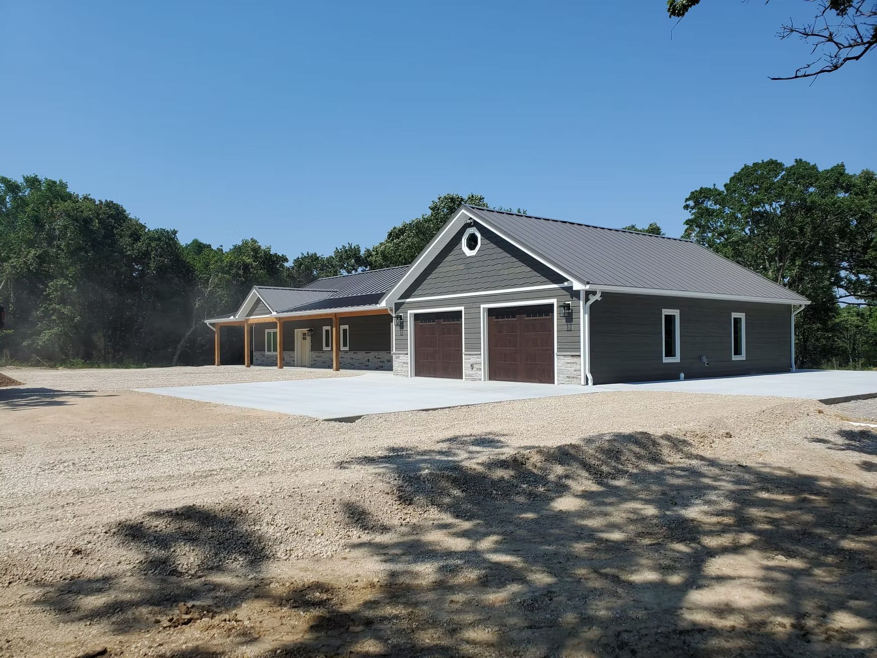 Tan metal shed with a brown roof and trim, set on a gravel base against a backdrop of green trees and a blue sky.