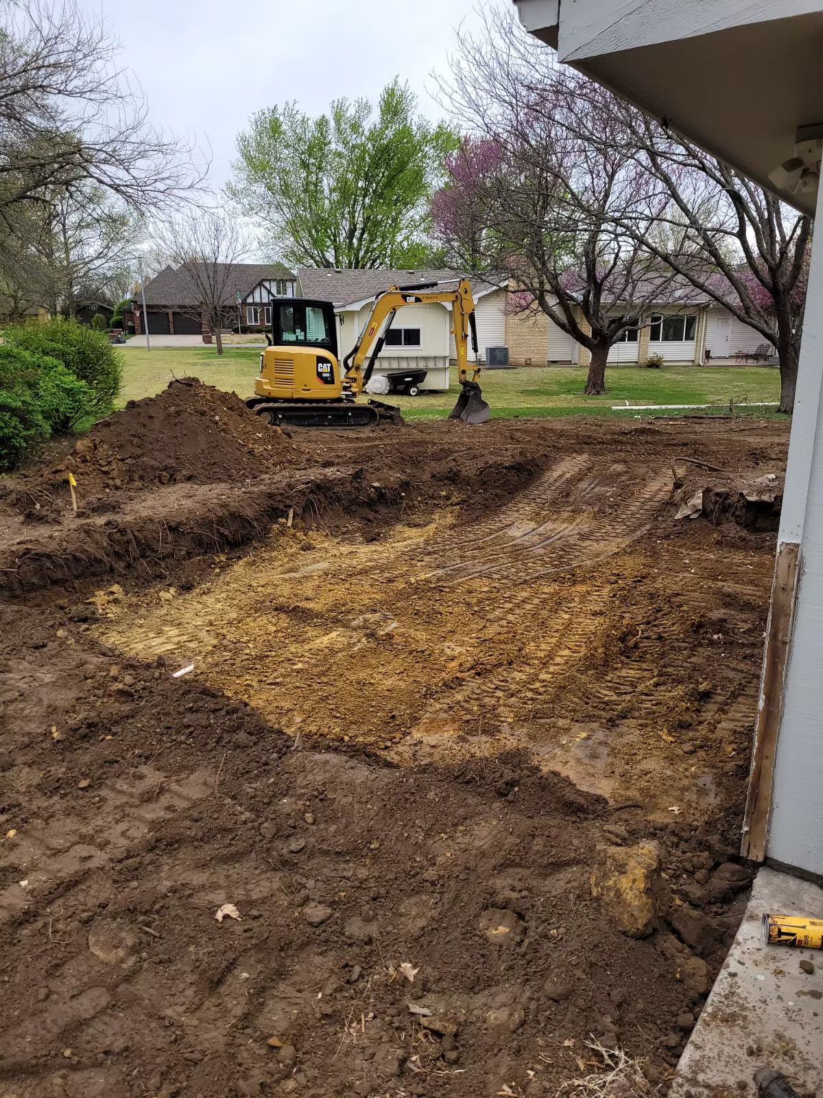 A small excavator digs in a yard, preparing the ground with piles of soil. Houses and trees are in the background.